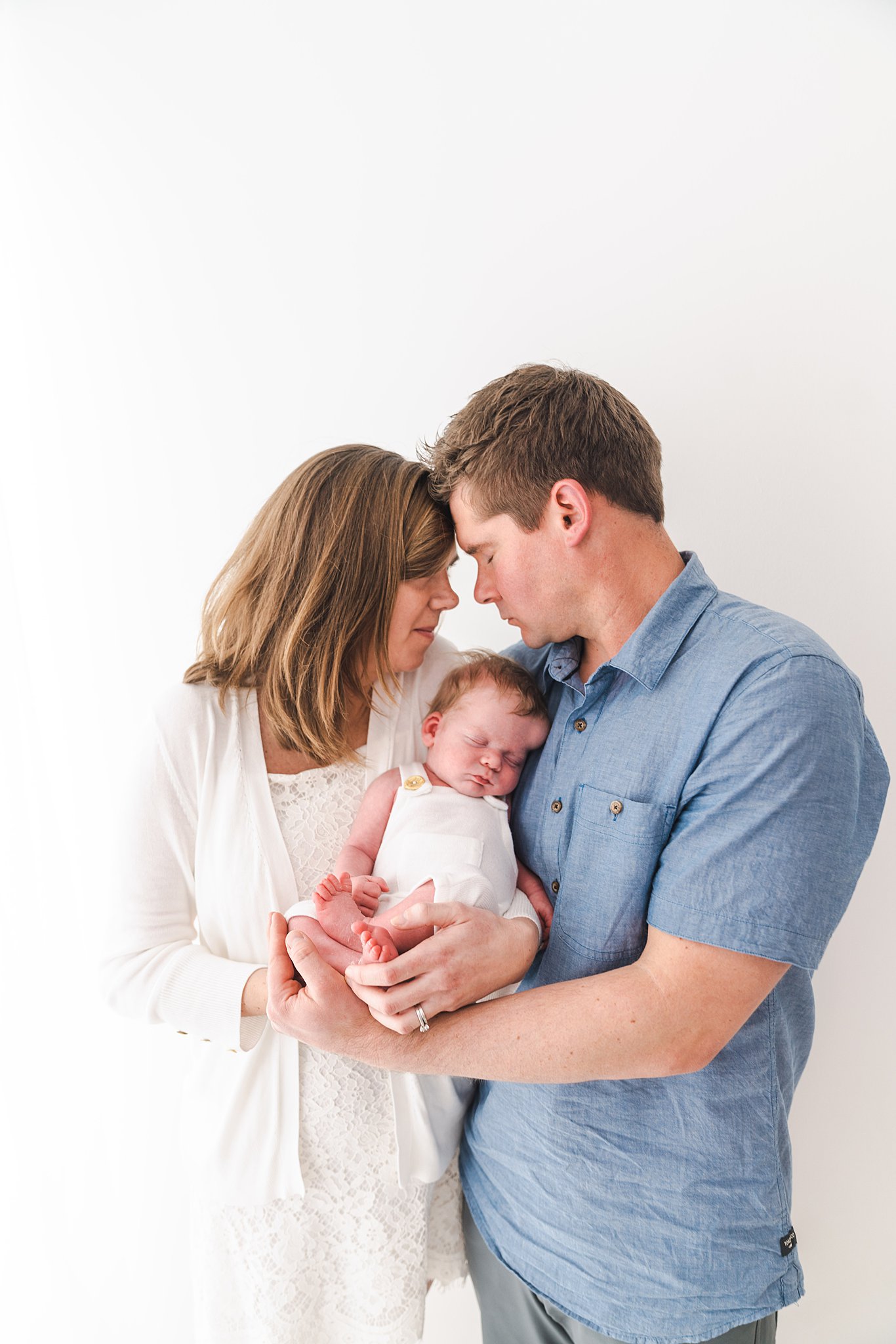 mother in white dress puts forehead against father in blue denim shirt while holding their sleeping newborn in a studio lactation consultant minneapolis