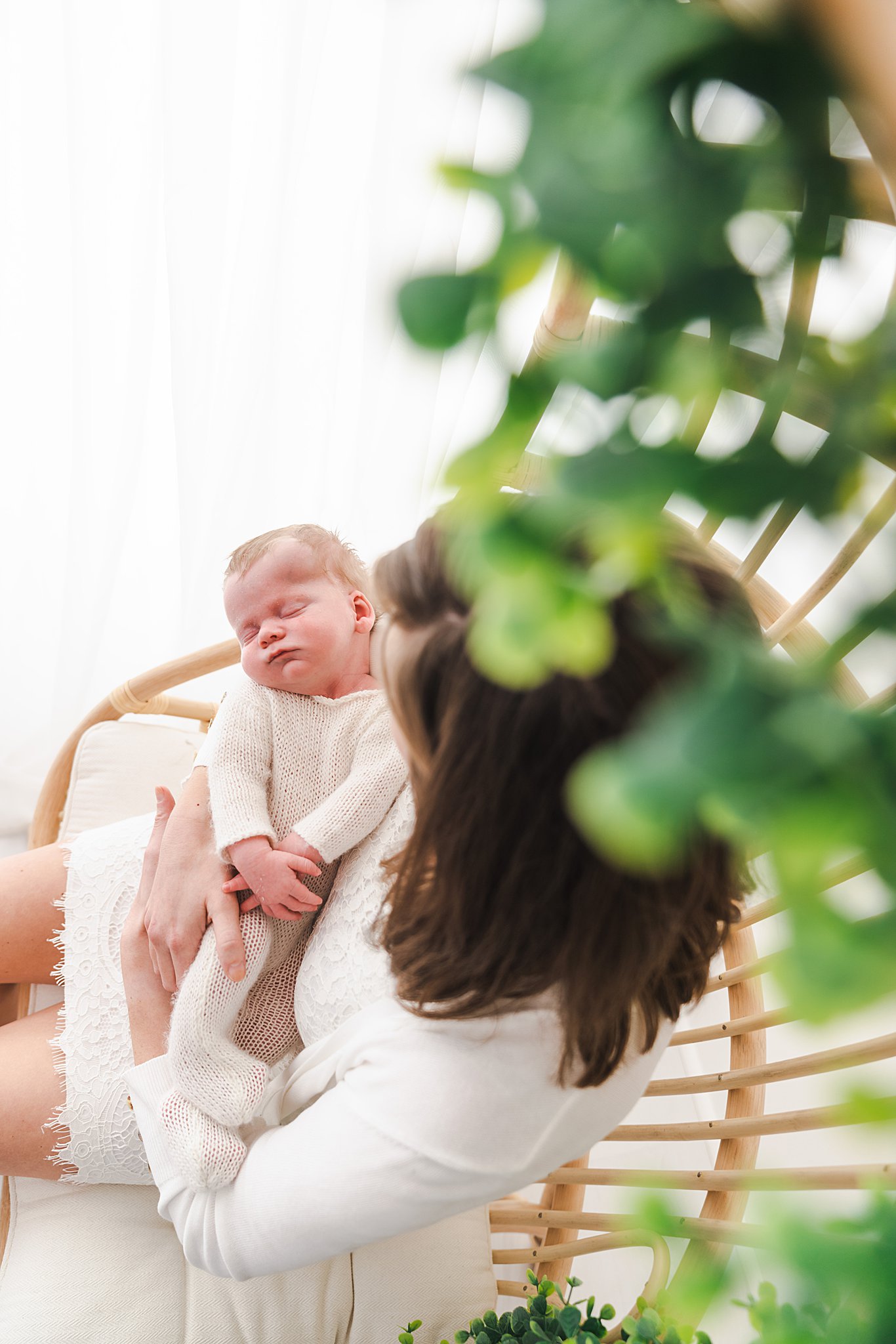 mother in white dress sits on a tall wicker chair holding her newborn while they sleep