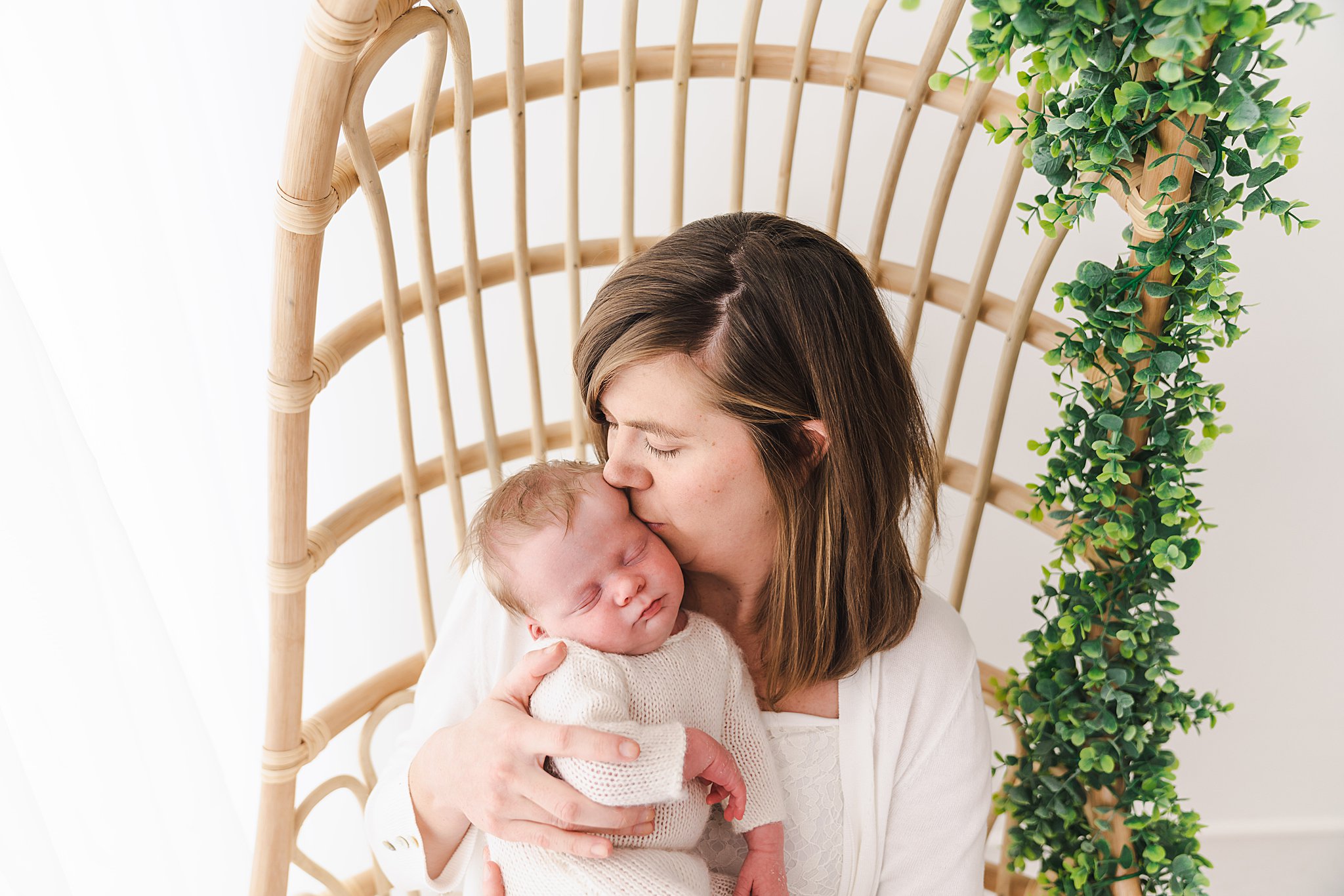 Mother in white blouse kisses the head of her newborn wearing a white knit onesie on a wicker chair in a studio lactation consultant minneapolis