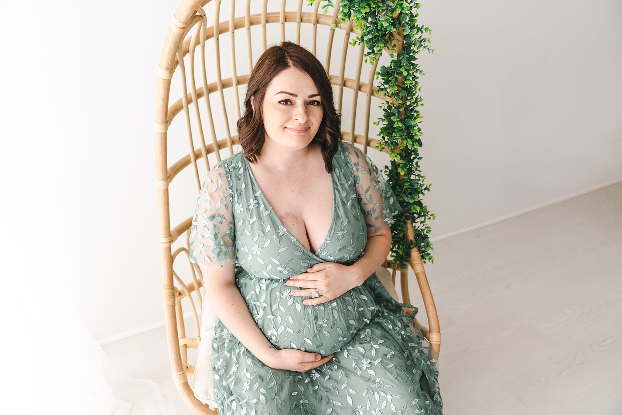 Mother to be wearing a green leaf patterned maternity gown sits in a studio in a wicker chair