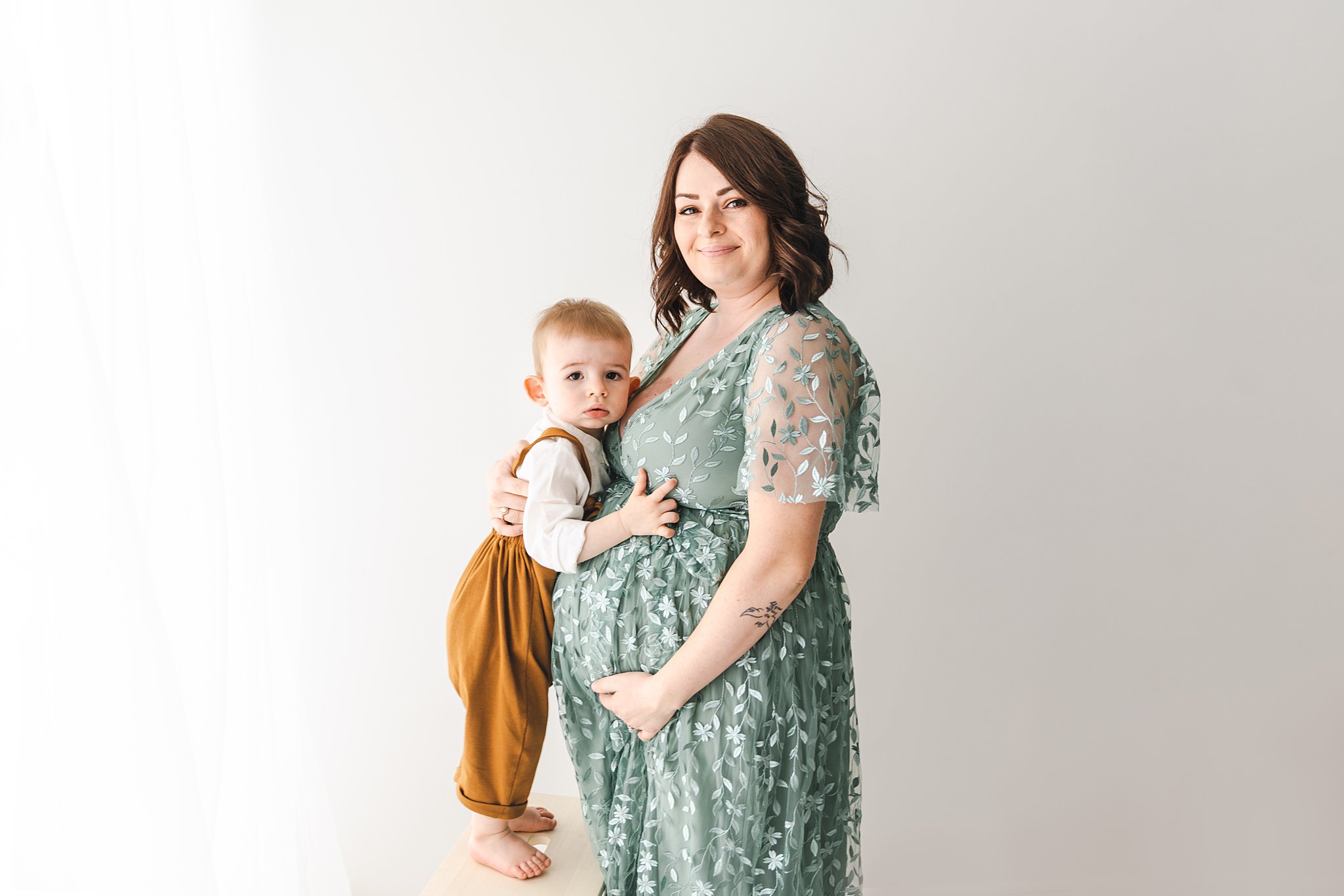 Mother wearing a green maternity gown holds her bump while son stands on a stool hugging her twin cities doulas
