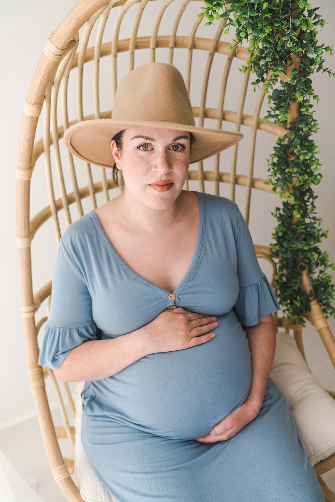Mother to be in a blue maternity dress sits in a wicker chair twin cities midwives