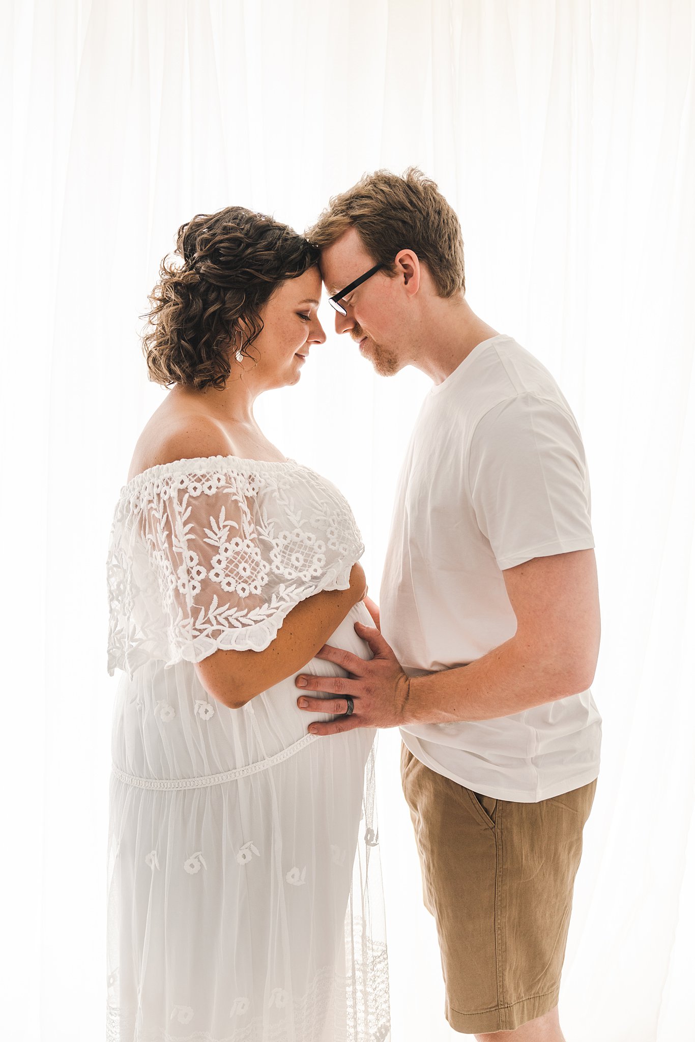 Mother to be stands forehead to forehead in a studio in front of a large window