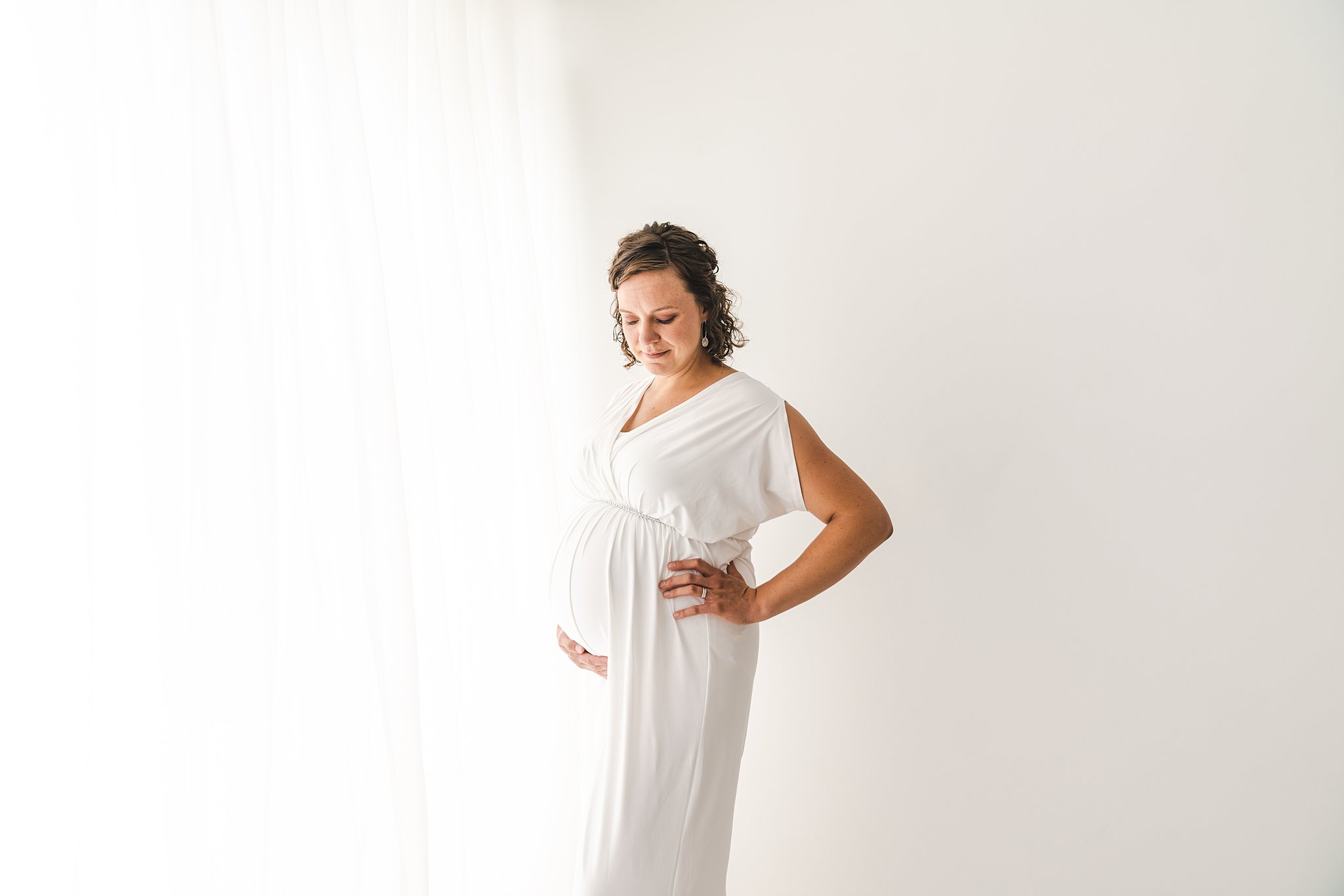 Mother to be stands in a studio next to a sarge window in a white maternity gown Blooma minneapolis