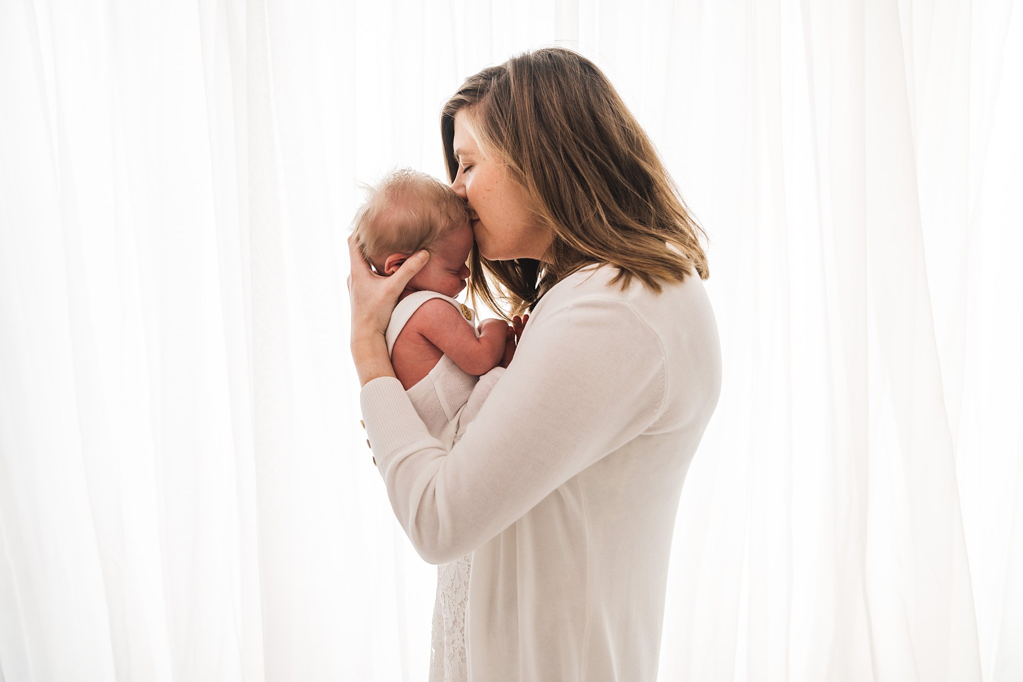 A mother in a white sweater stands in front of a window holding and kissing the forehead of her newborn baby Welcome Baby Care