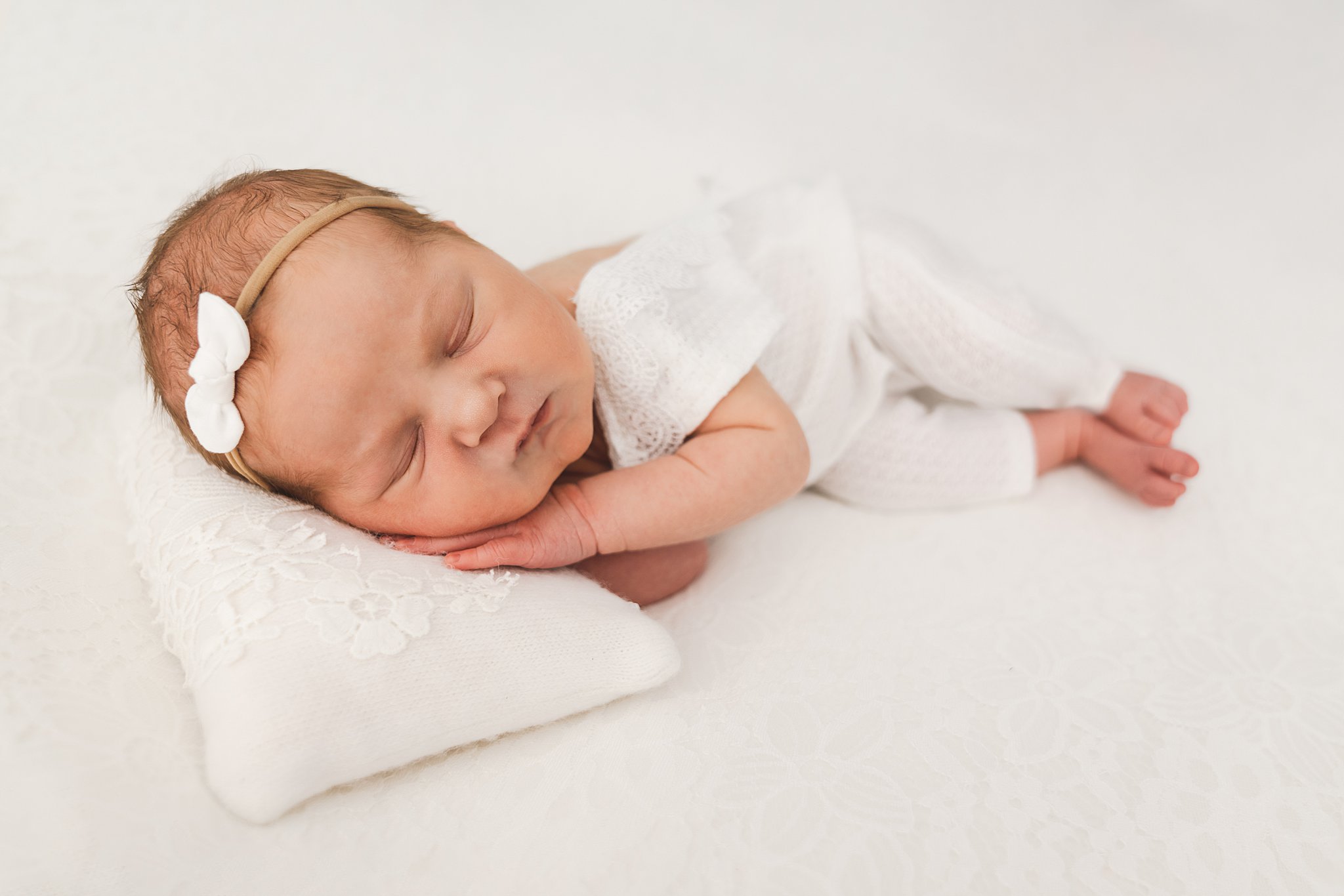 Newborn baby sleeps in a white onesie with hands under head on a pillow in a studio