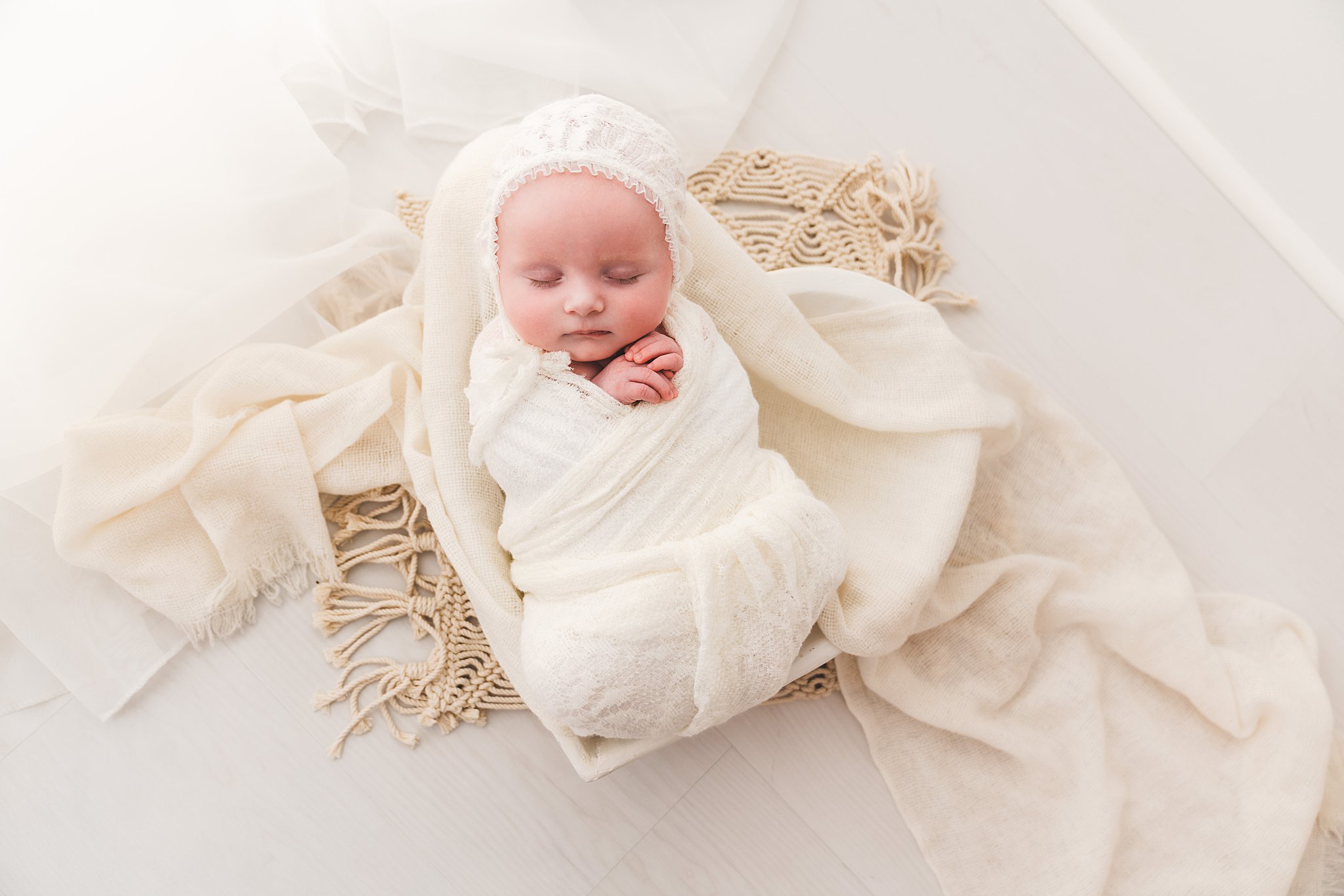A sleeping newborn baby sleeps in a studio with hands folded in a white swaddle and bonnet