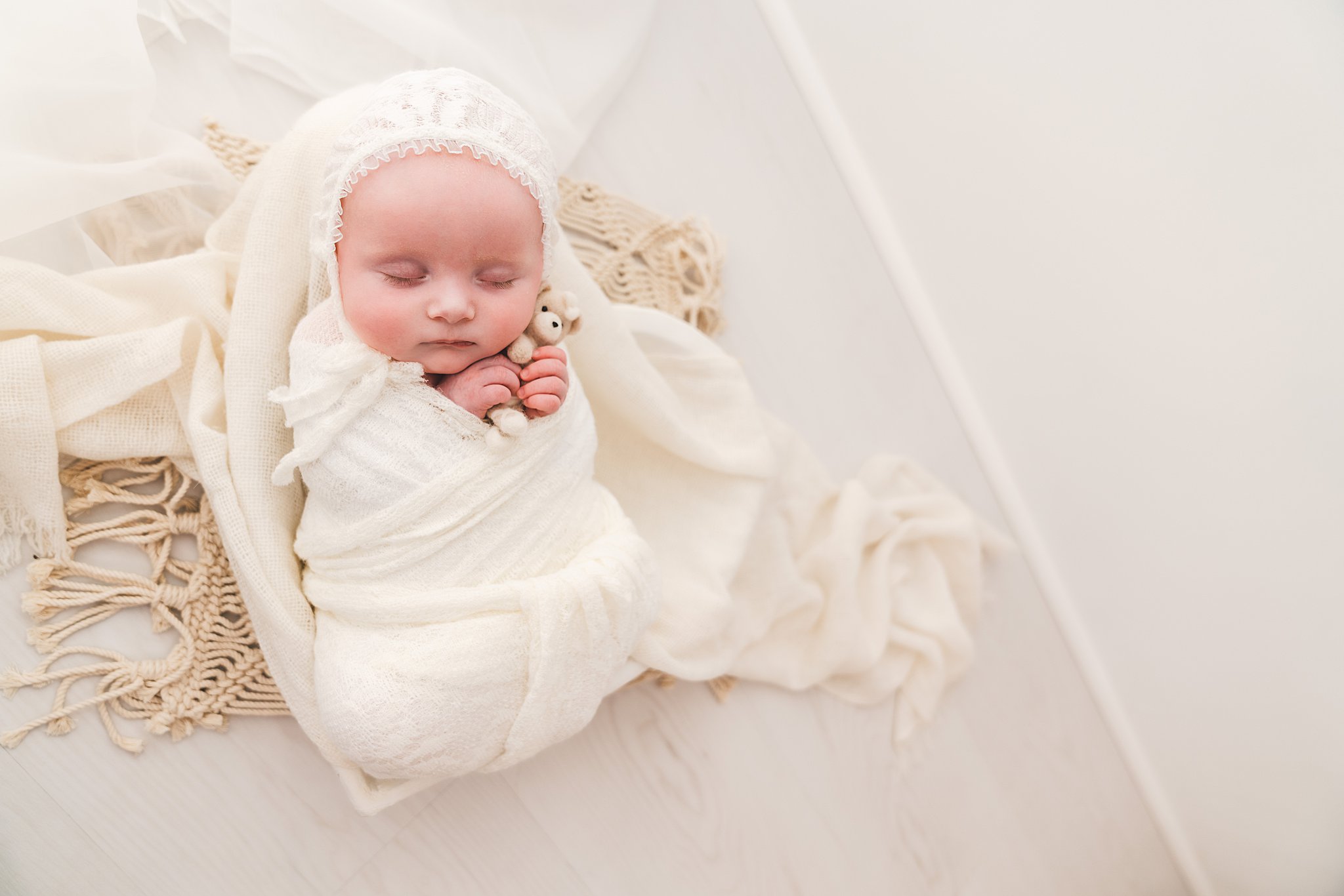 Newborn baby sleeps and cuddles a small stuffed bear in a lace bonnet and white swaddle oh baby minneapolis