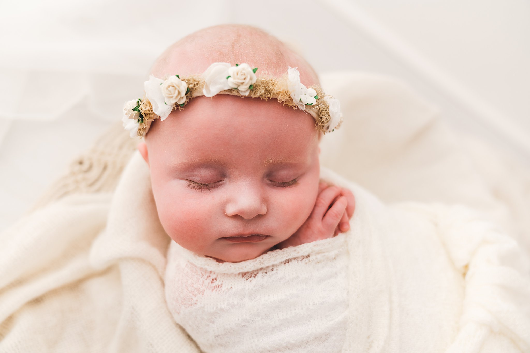 Newborn baby sleeps in a white swaddle on a bed of blankets in a floral headband oh baby minneapolis