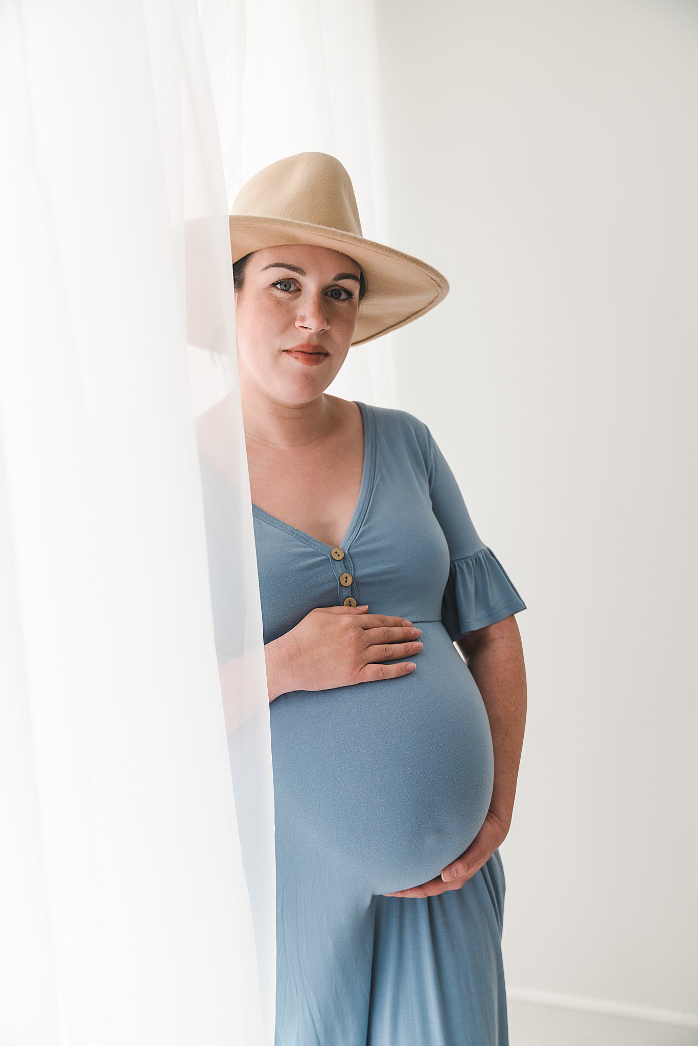 A mother to be in a blue maternity gown and tan hat stands in the white curtains of a window roots community birth center