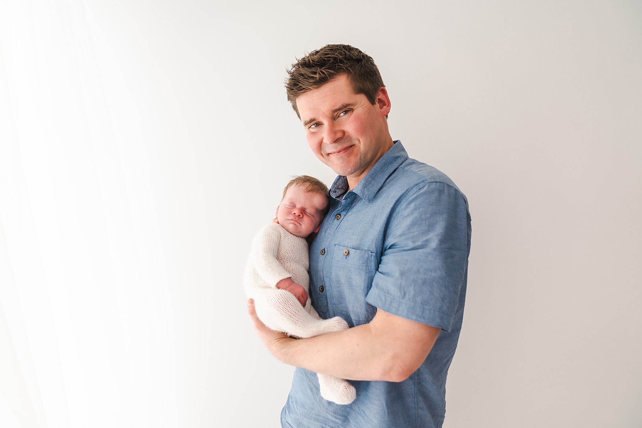 A proud father in a blue button down shirt stands in a studio holding his newborn baby in a white knit onesie roots community birth center