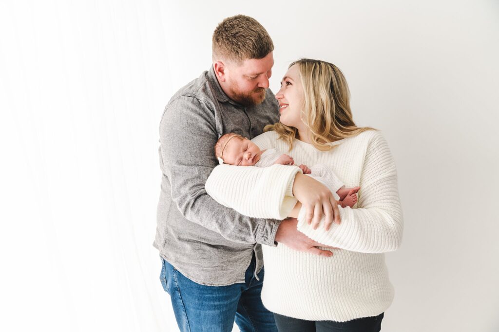 New dad in a grey shirt hugs onto his wife holding their sleeping newborn baby in a studio twin cities midwifery