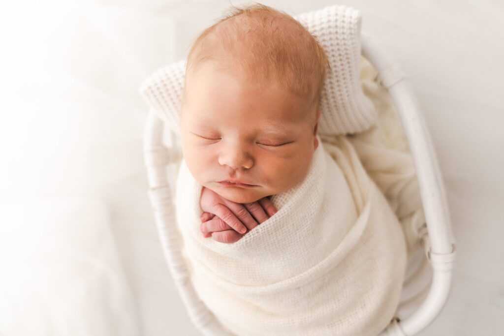 A newborn baby sleeps in a white swaddle in a white wicker basket baby grand minnesota