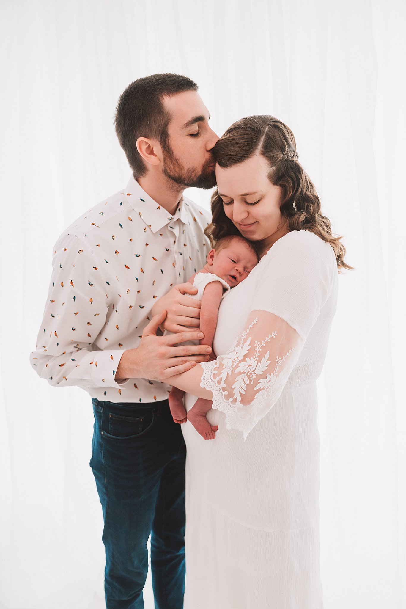 A new father kisses the forehead of his wife who is snuggling their sleeping newborn baby bumbelou