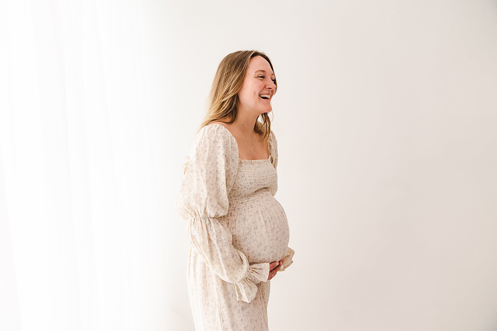 A mother to be laughs while standing with hands under her bump in a studio