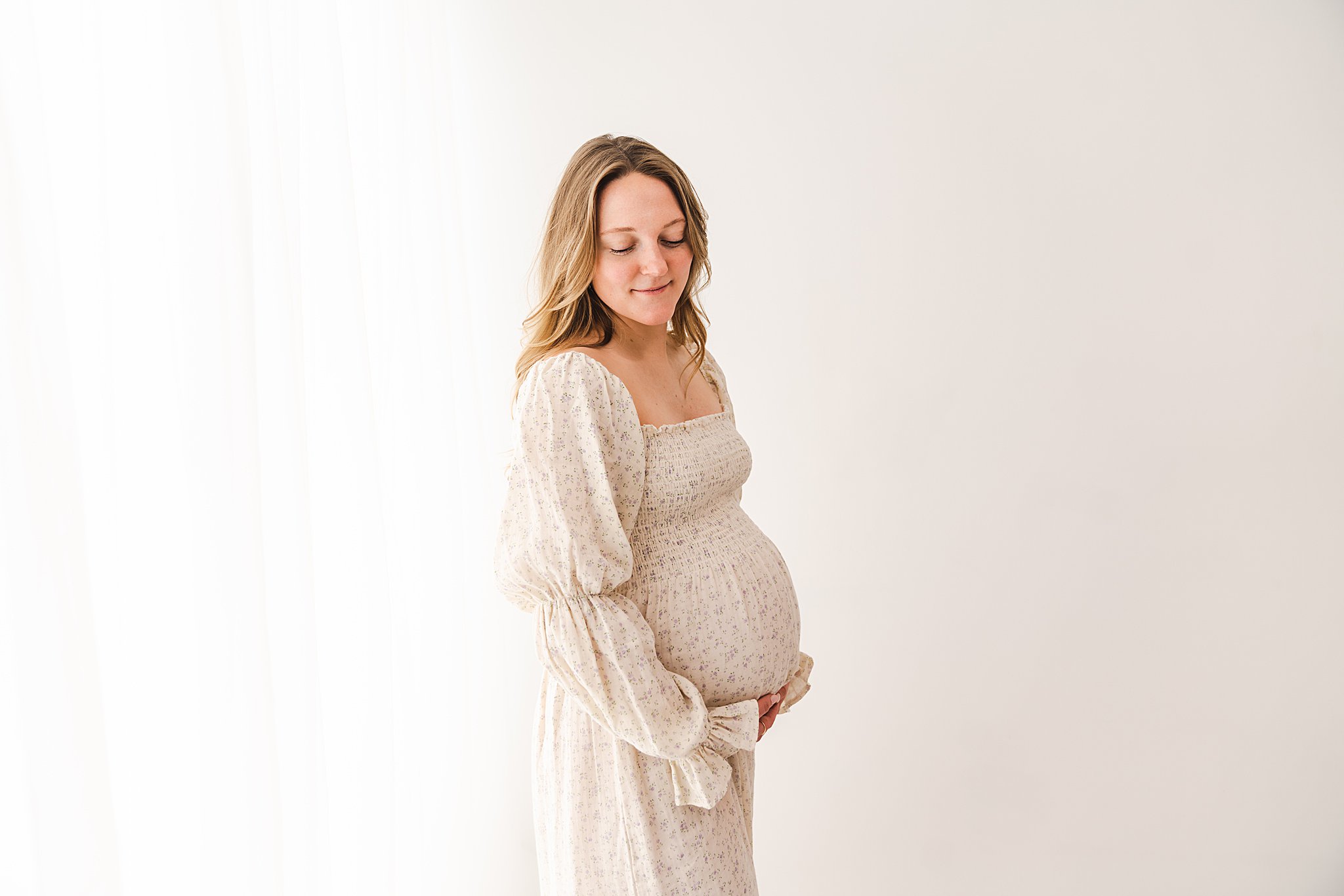 A mom to be stands in a studio looking down at her bump in a white maternity dress flutterby birth services