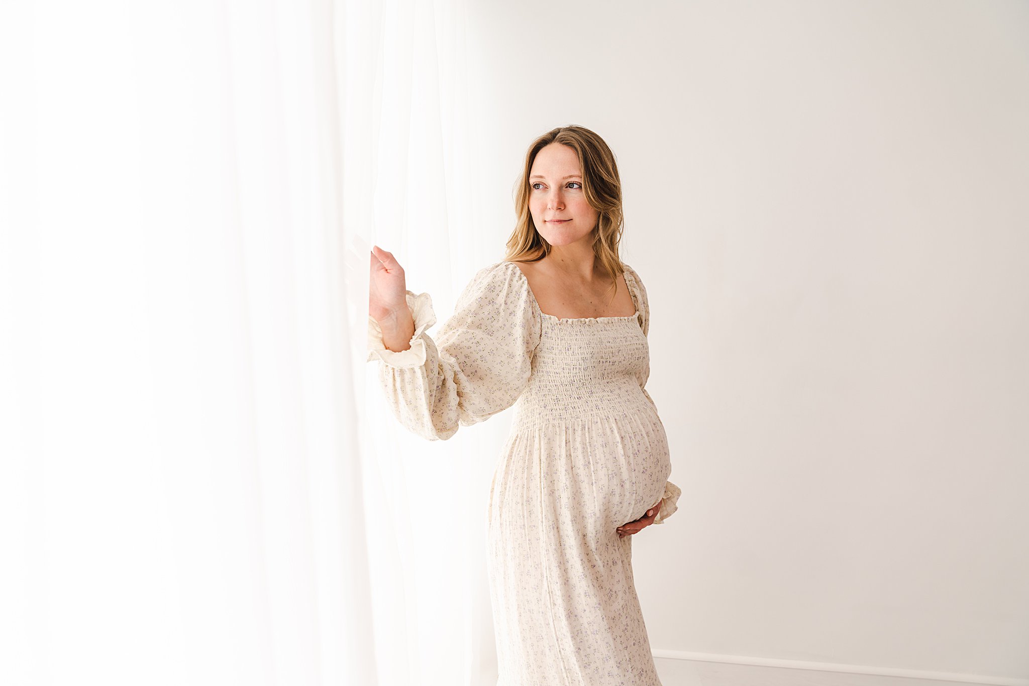 A mother to be stands in a window of a studio holding her bump and looking out thee window in a white maternity dress flutterby birth services