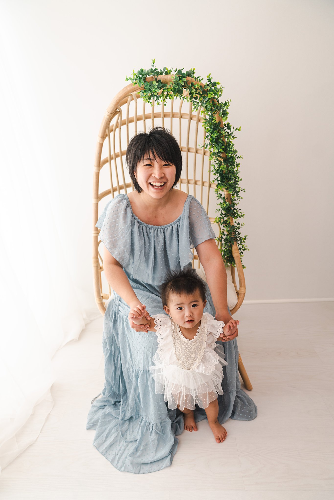 A mother in a blue dress helps her toddler stand while sitting in a tall wicker chair in a studio goodnight moon edina