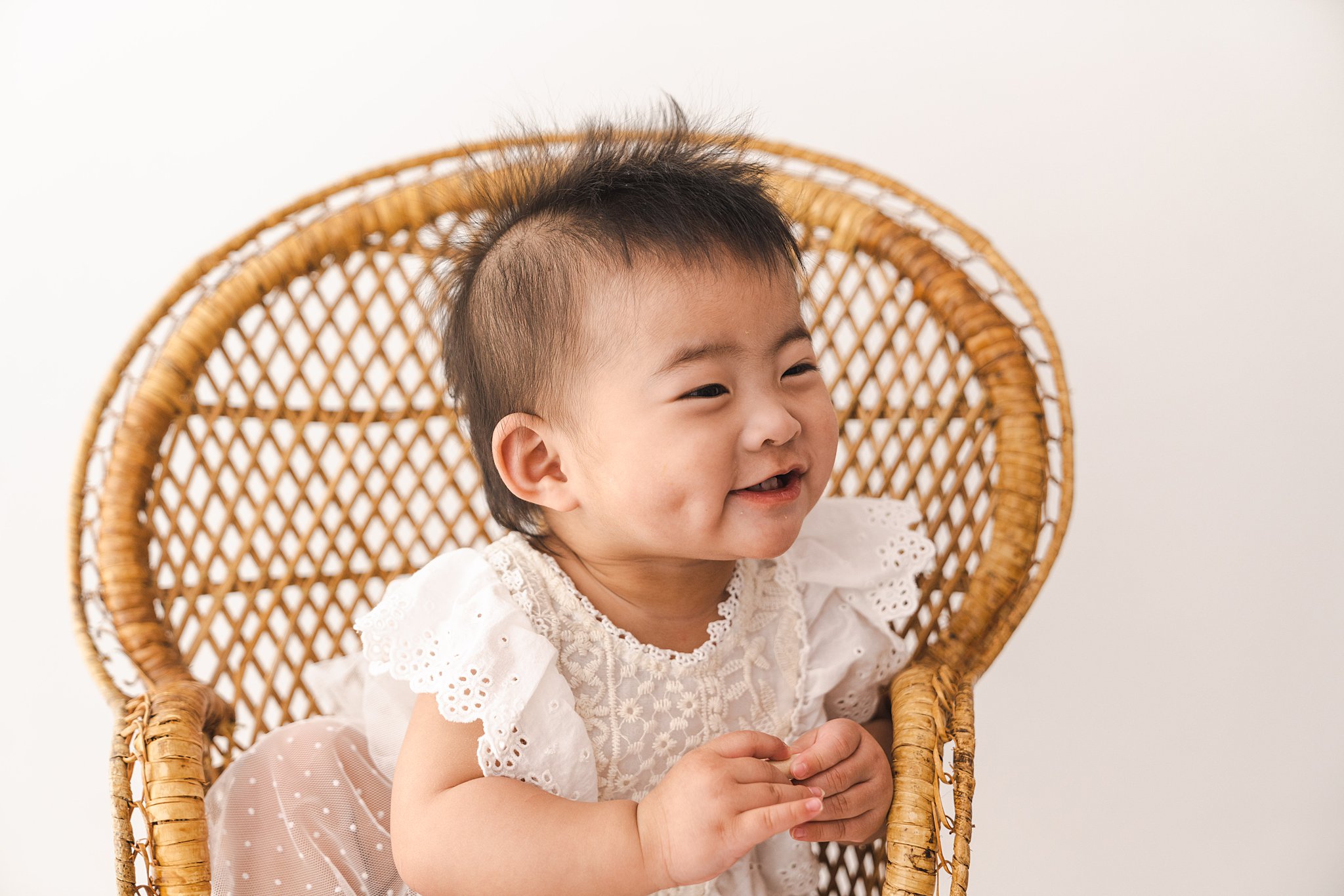 A young toddler in a white lace dress sits in a wicker chair in a studio goodnight moon edina