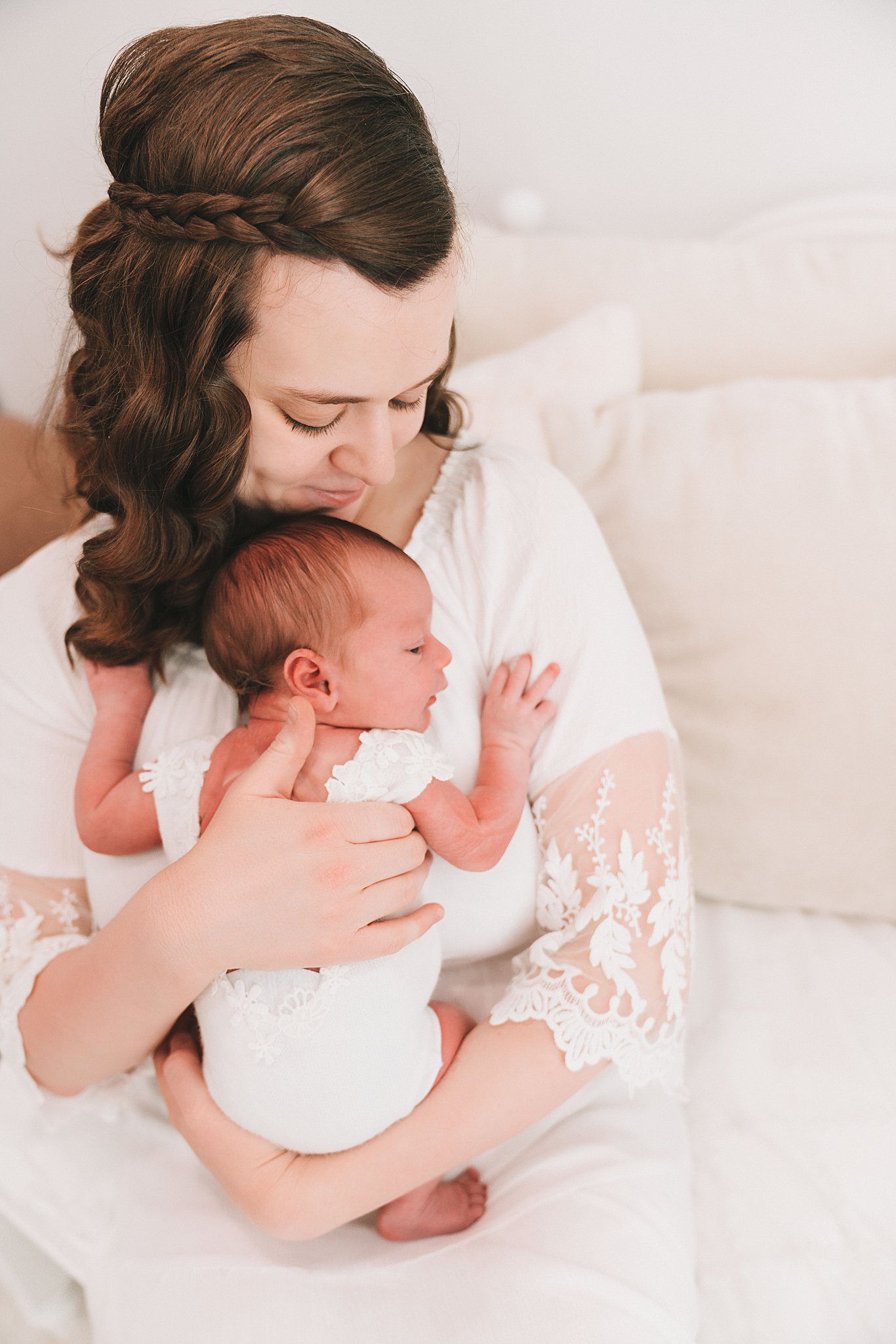 A mother in a white dress and lace sleeves sits on a couch holding her newborn baby against her chest