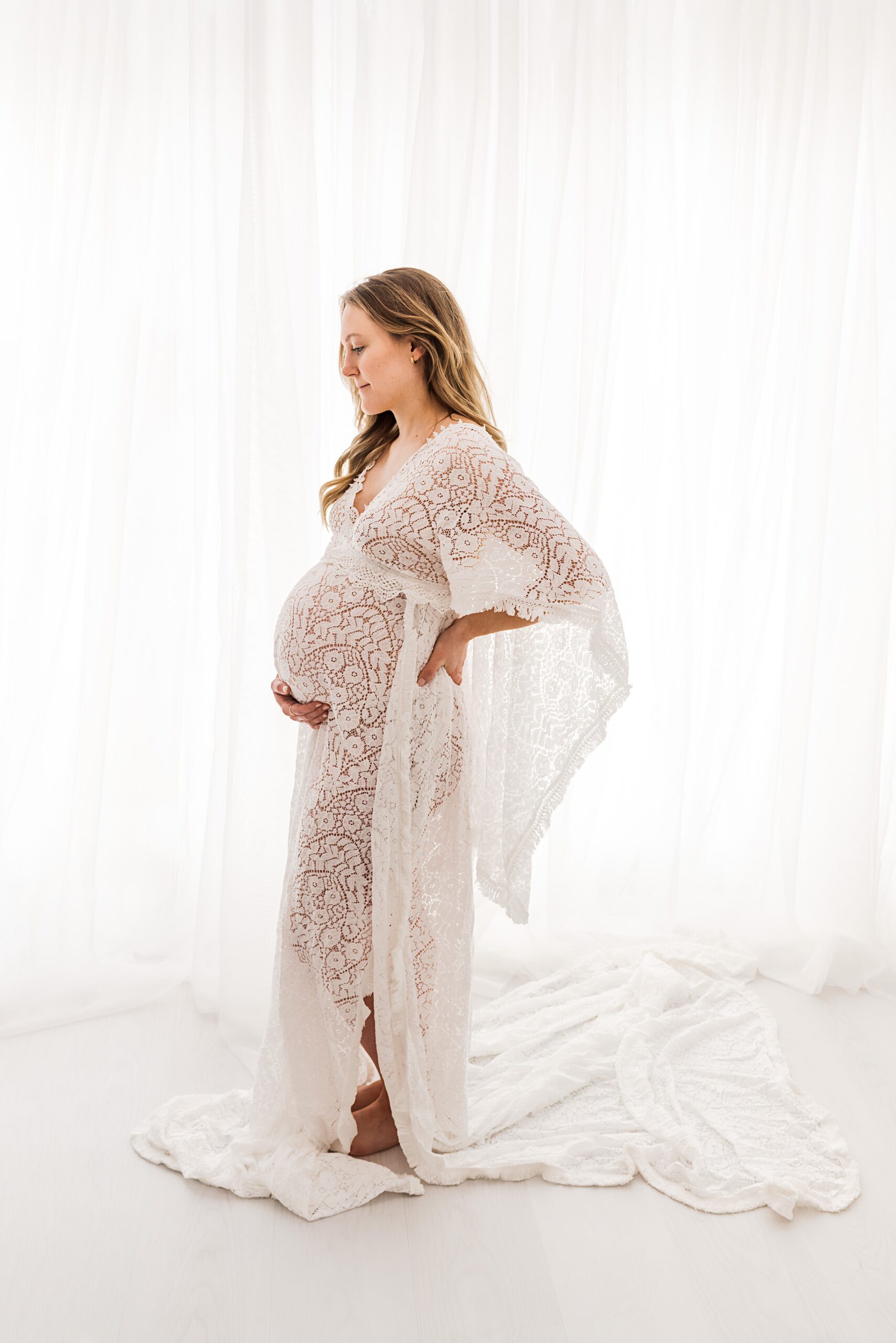 A mother to be in a long flowing lace maternity gown stands in front of a large window holding her bump mother baby center minneapolis