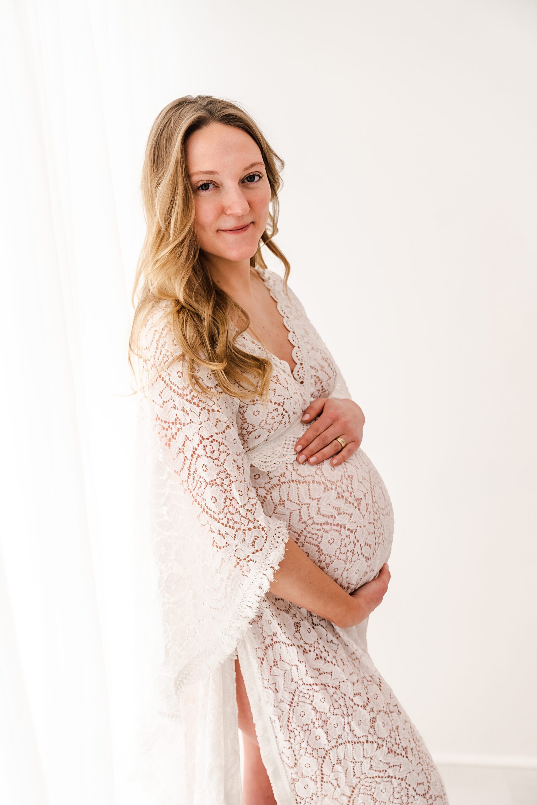 A mom to be in a lace maternity gown stands in a studio by a window mother baby center minneapolis