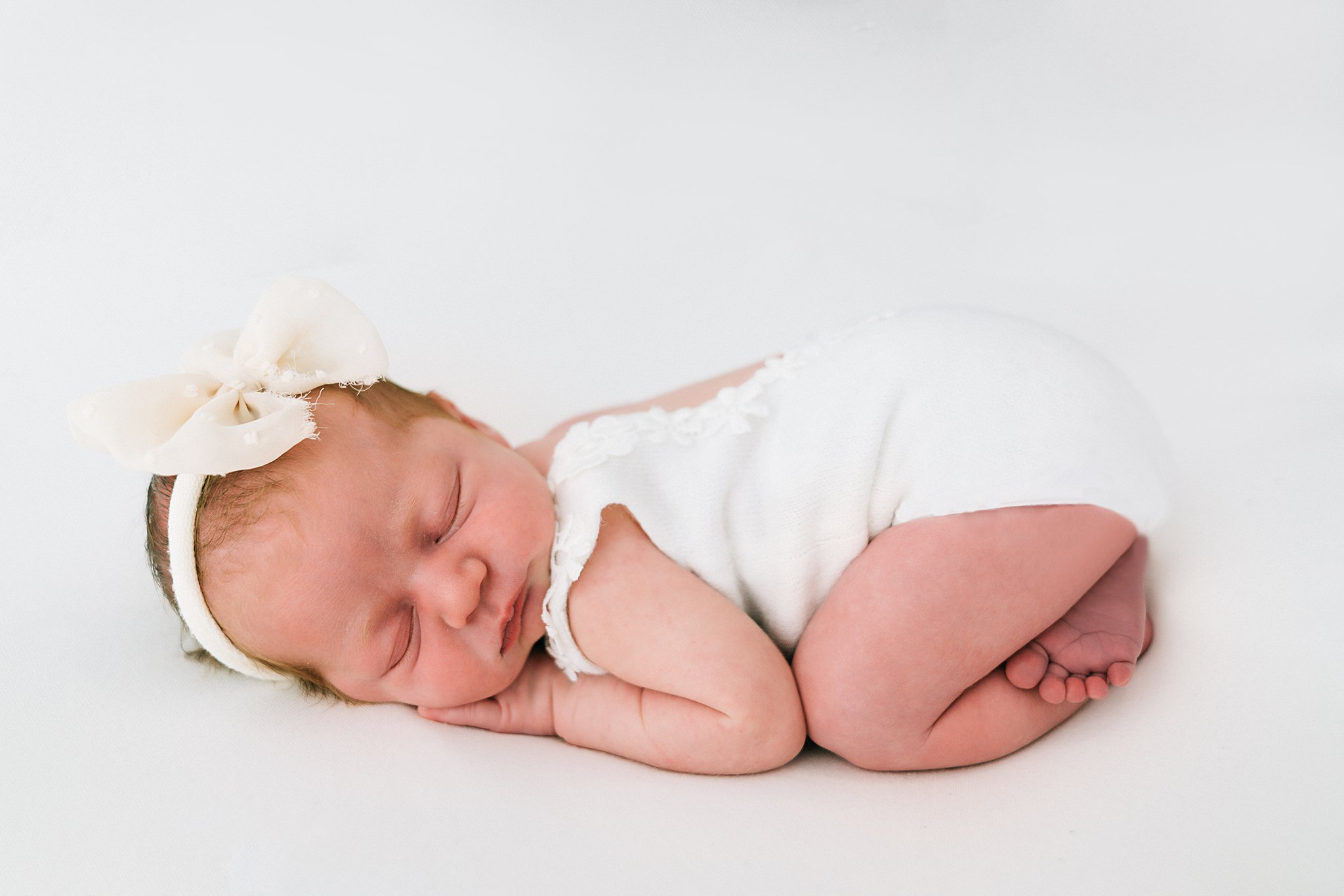 A newborn baby sleeps in froggy pose in a white onesie and bow headband