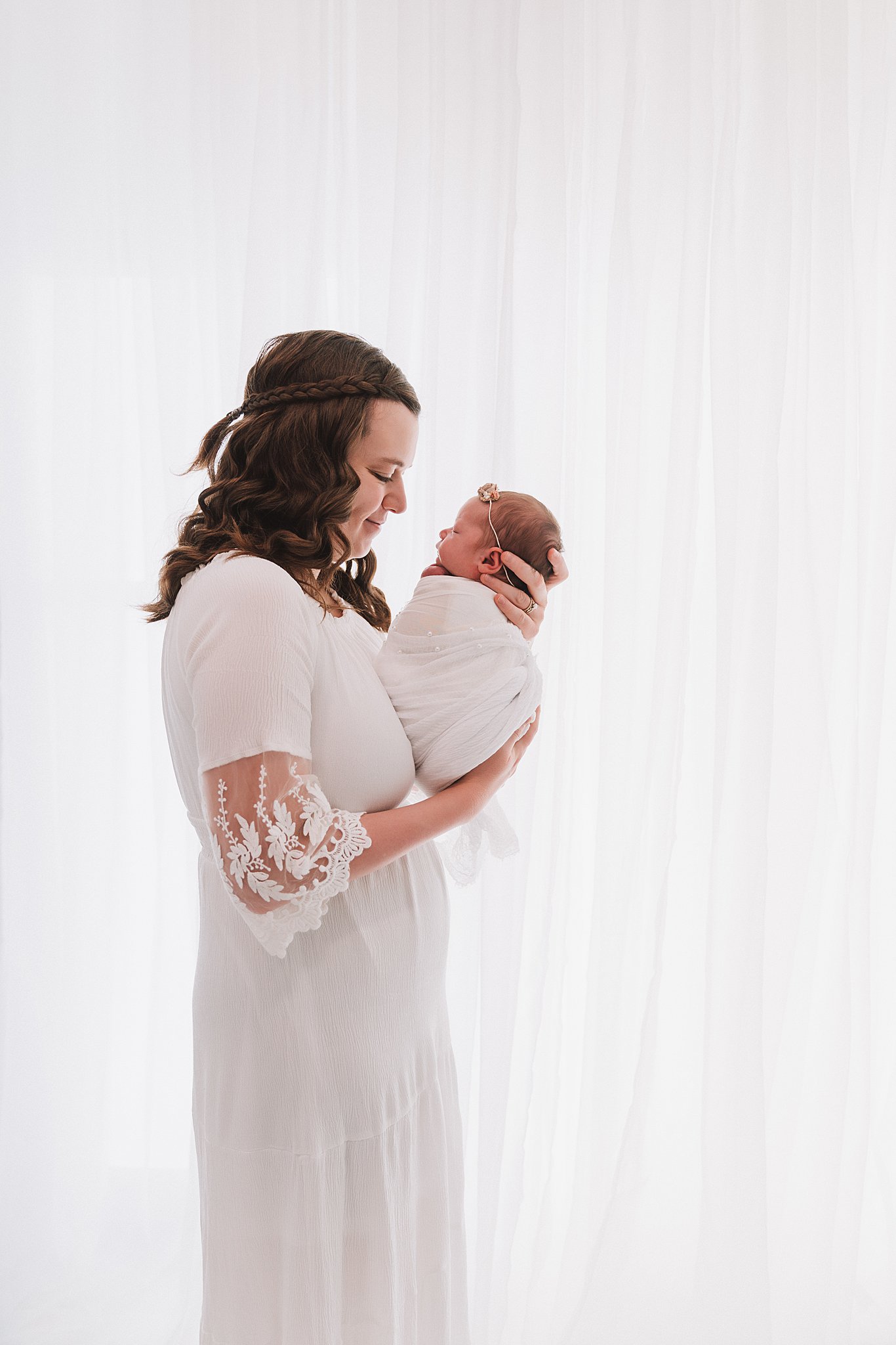 A mother in a white dress stands in front of a large window holding her newborn baby in a white swaddle pacifier minneapolis