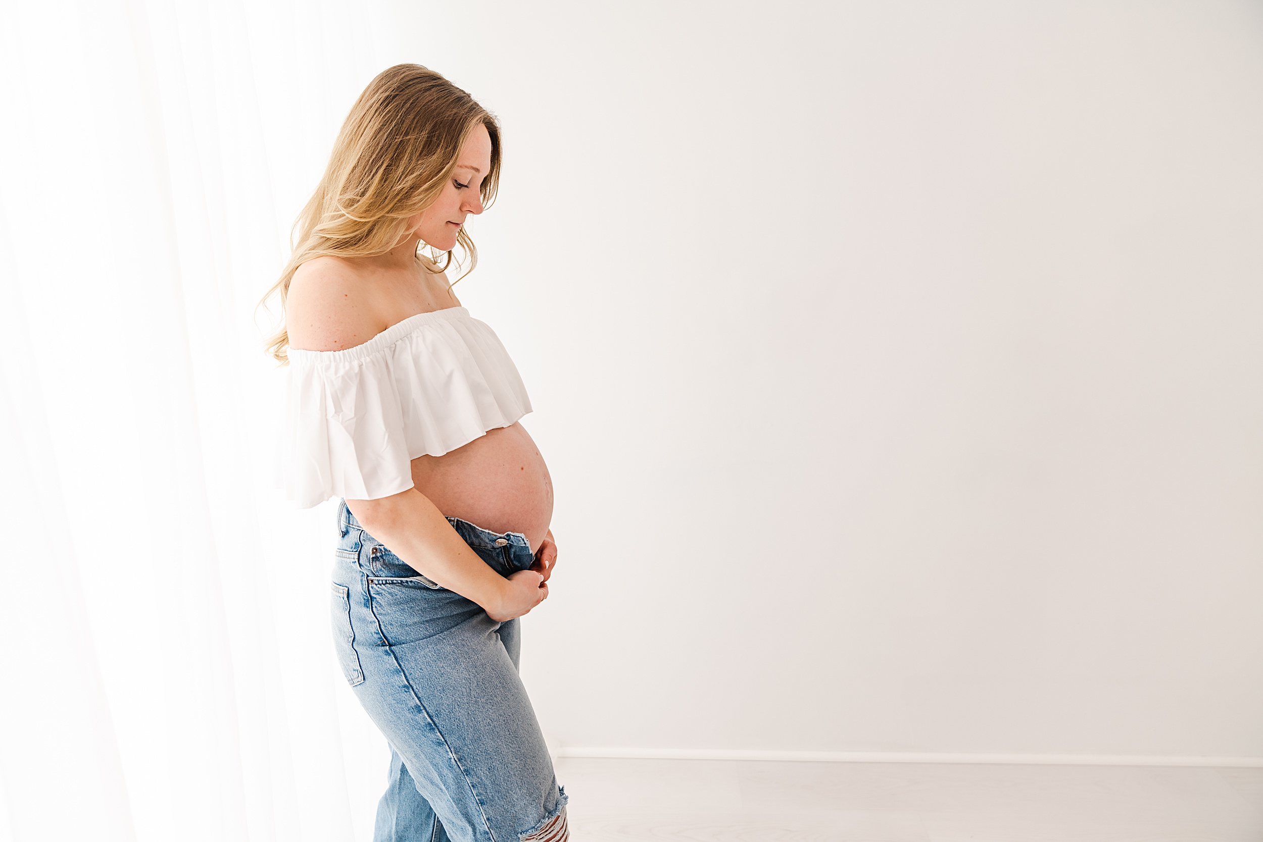 A mom to be stands in a studio by a window in ripped jeans looking down and holding the bottom of her bump