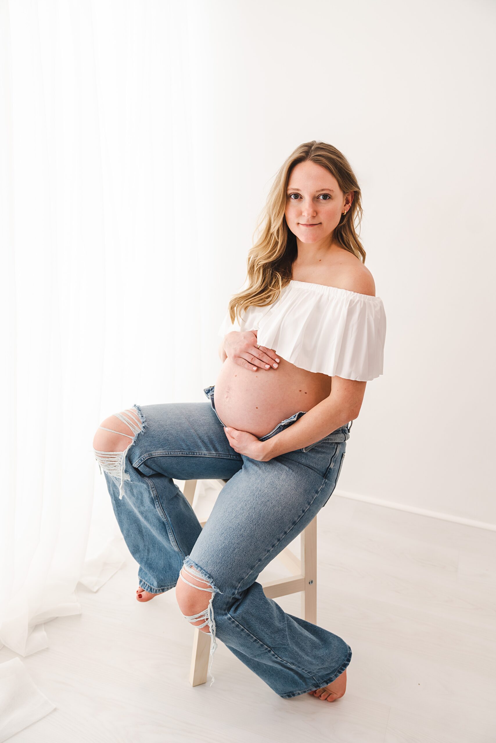 A mother to be in ripped jeans sits on a stool holding her bump in a studio thrive midwives