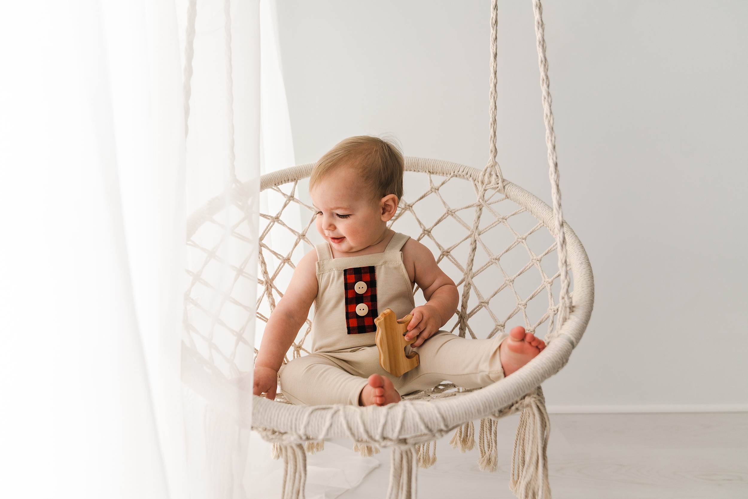 A young toddler in tan overalls sits in a rope swing hammock playing with a wooden bear kiddywampus