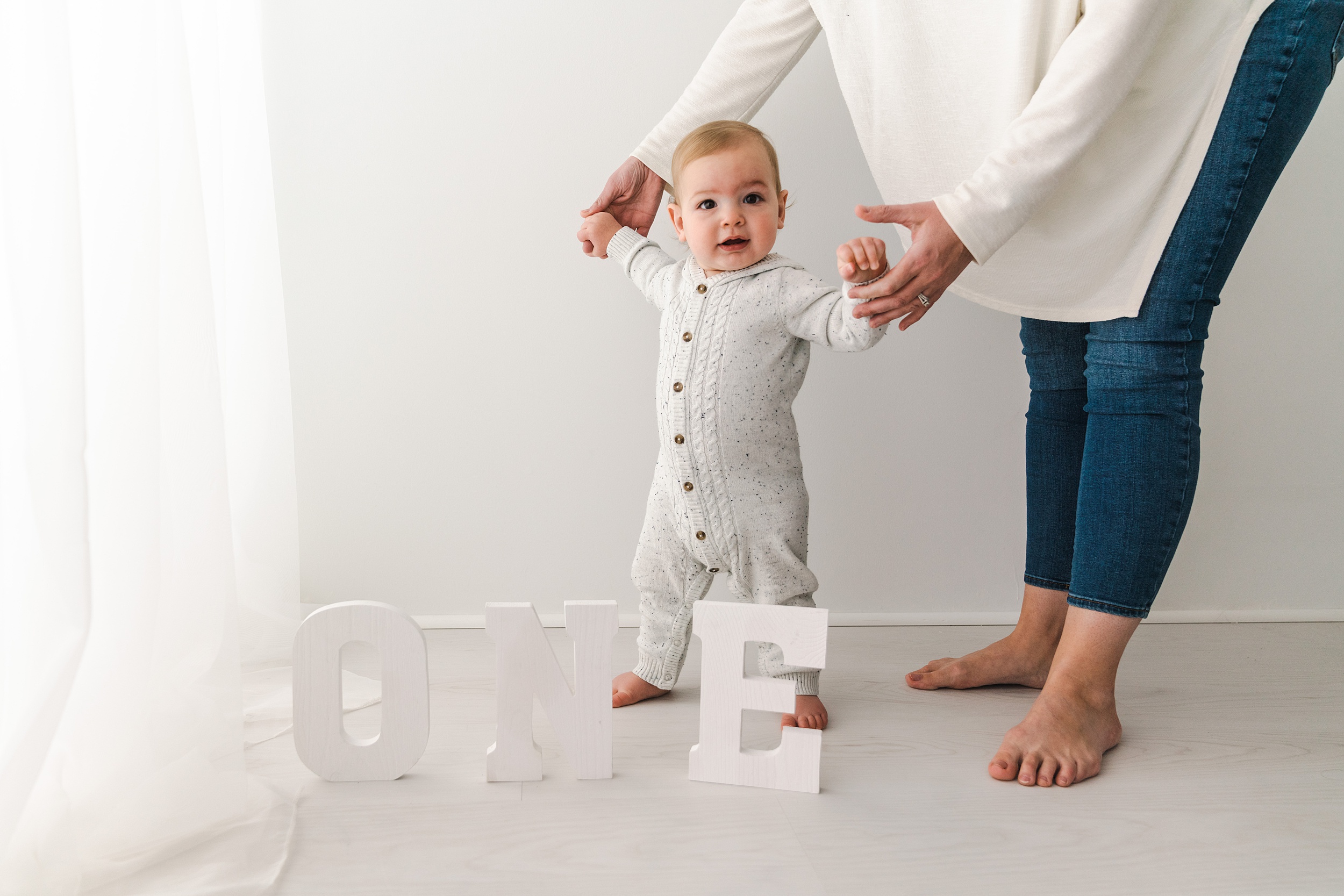 A mother holds the hands of her standing one year old in a studio