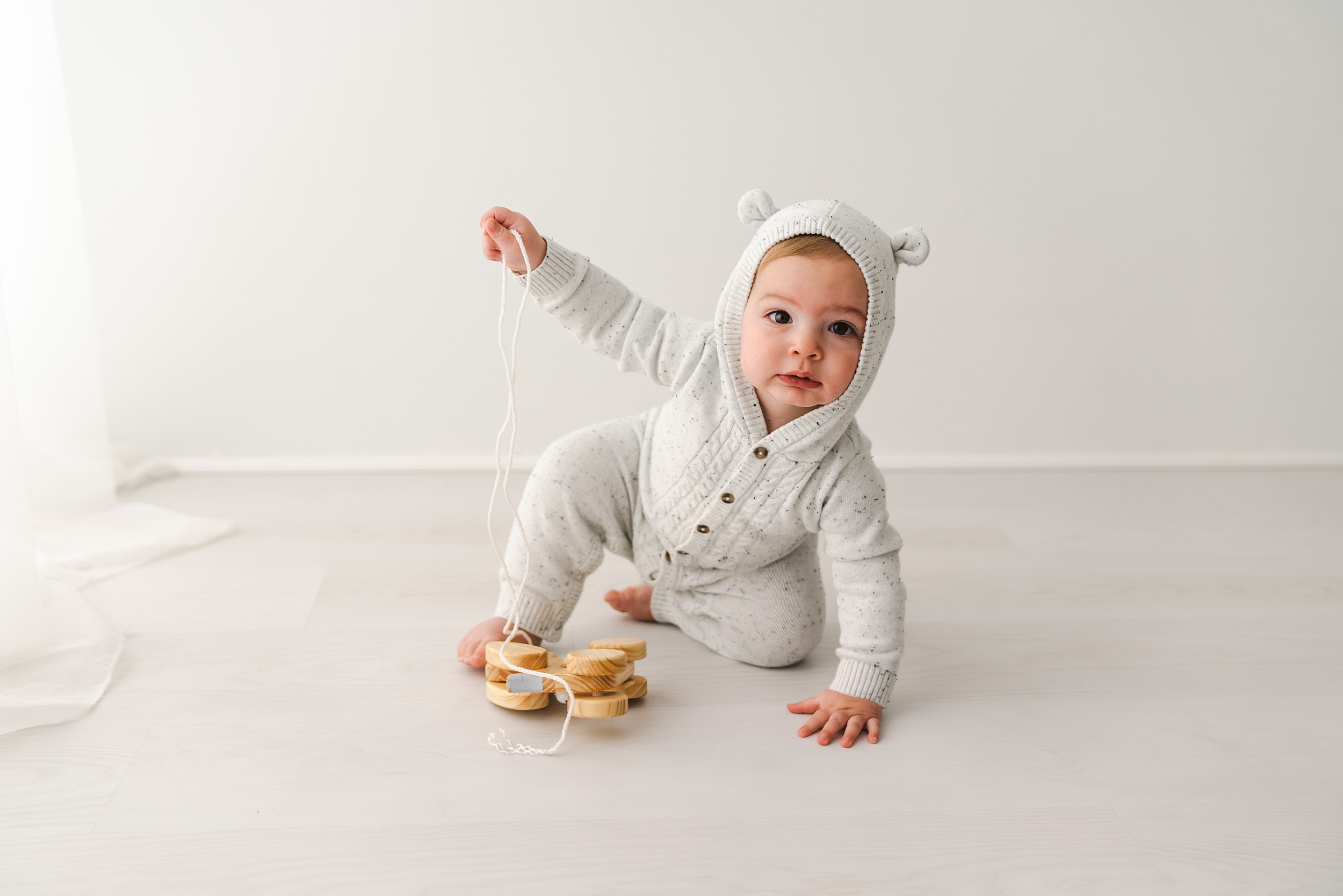 A toddler boy plays on the floor of a studio with a wooden car on a string while wearing a white onesie with ears kiddywampus