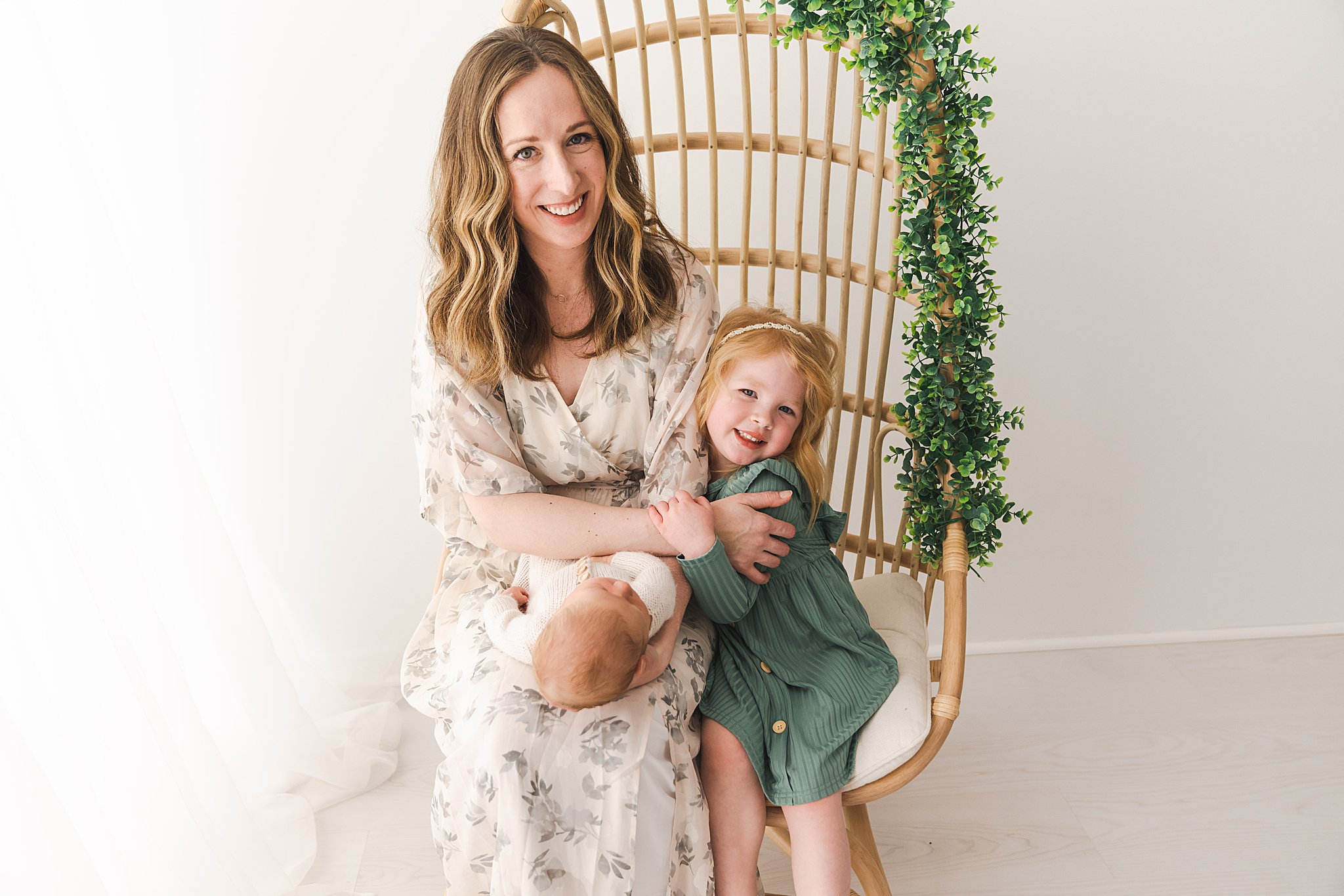 A mother sits on a tall wicker chair with her newborn baby in her lap and older daughter by her side moon and stars boutique