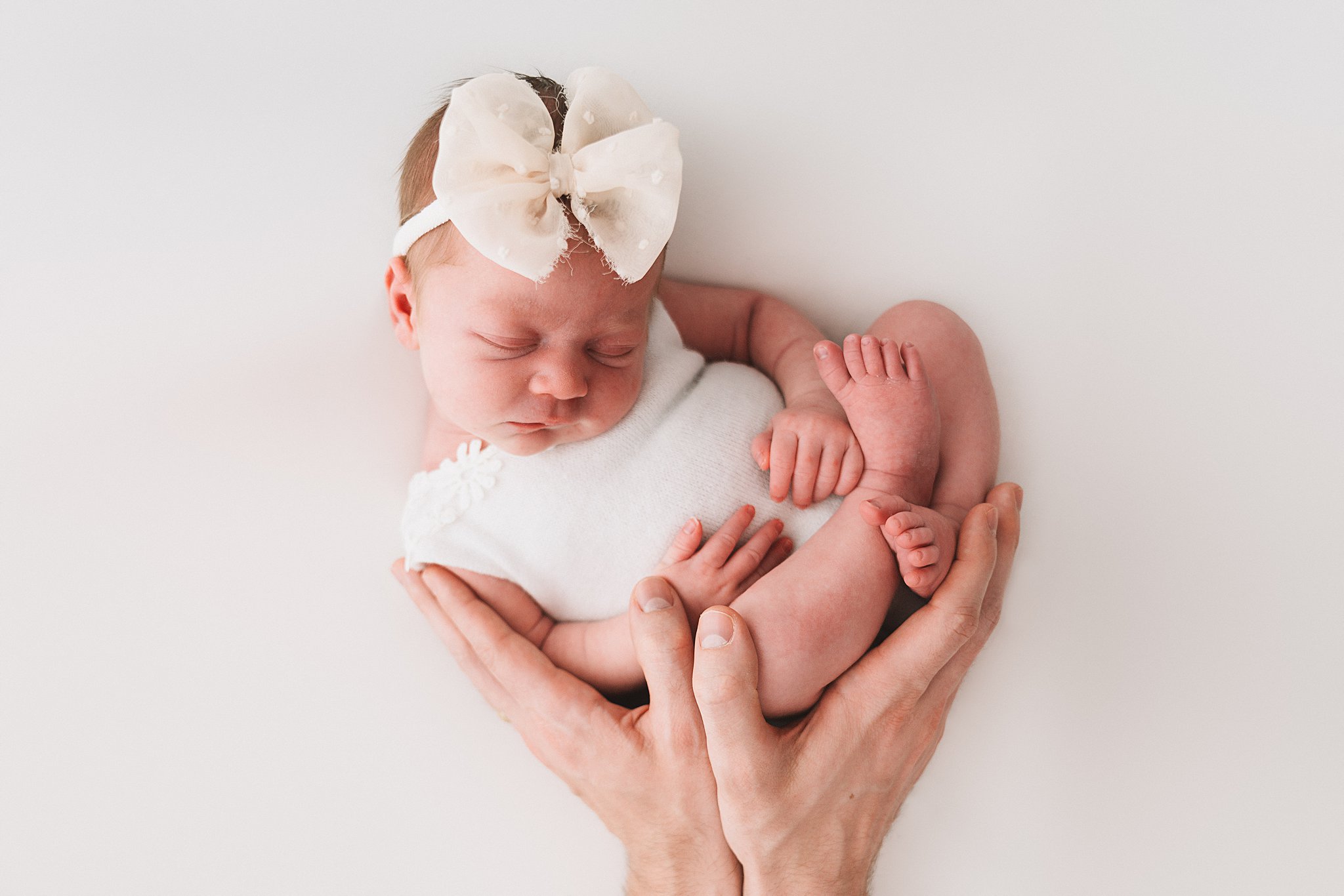 A newborn baby sleeps in a white onesie while being held by a pair of hands under her night nurse minneapolis