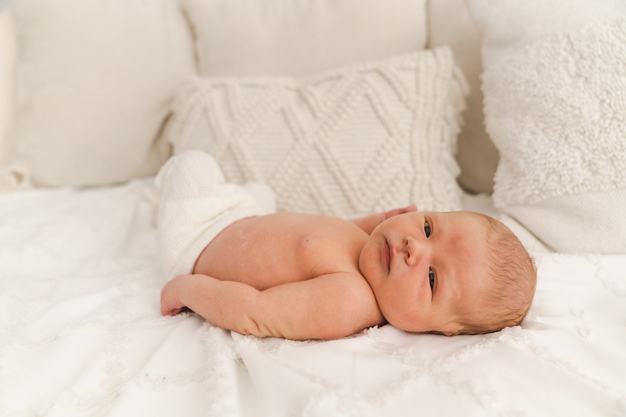 A newborn baby lays on a bed wearing white knit pants with eyes open