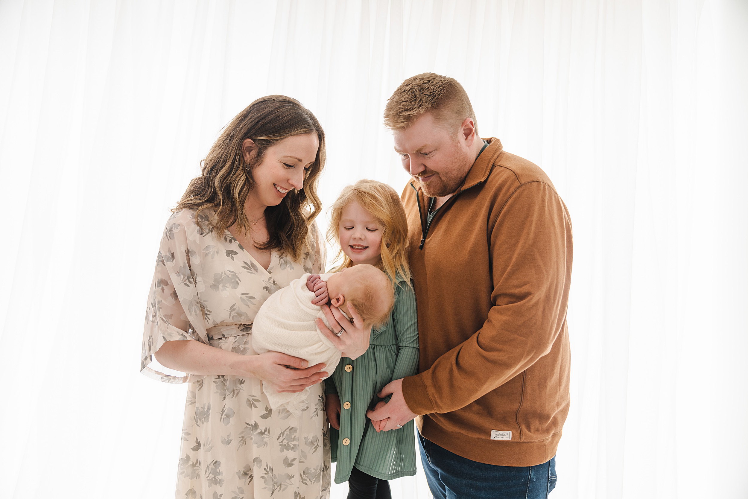 A mother and father stand with with their toddler daughter while looking down at the newborn baby in mom's arms in front of a window in a studio pip and pal