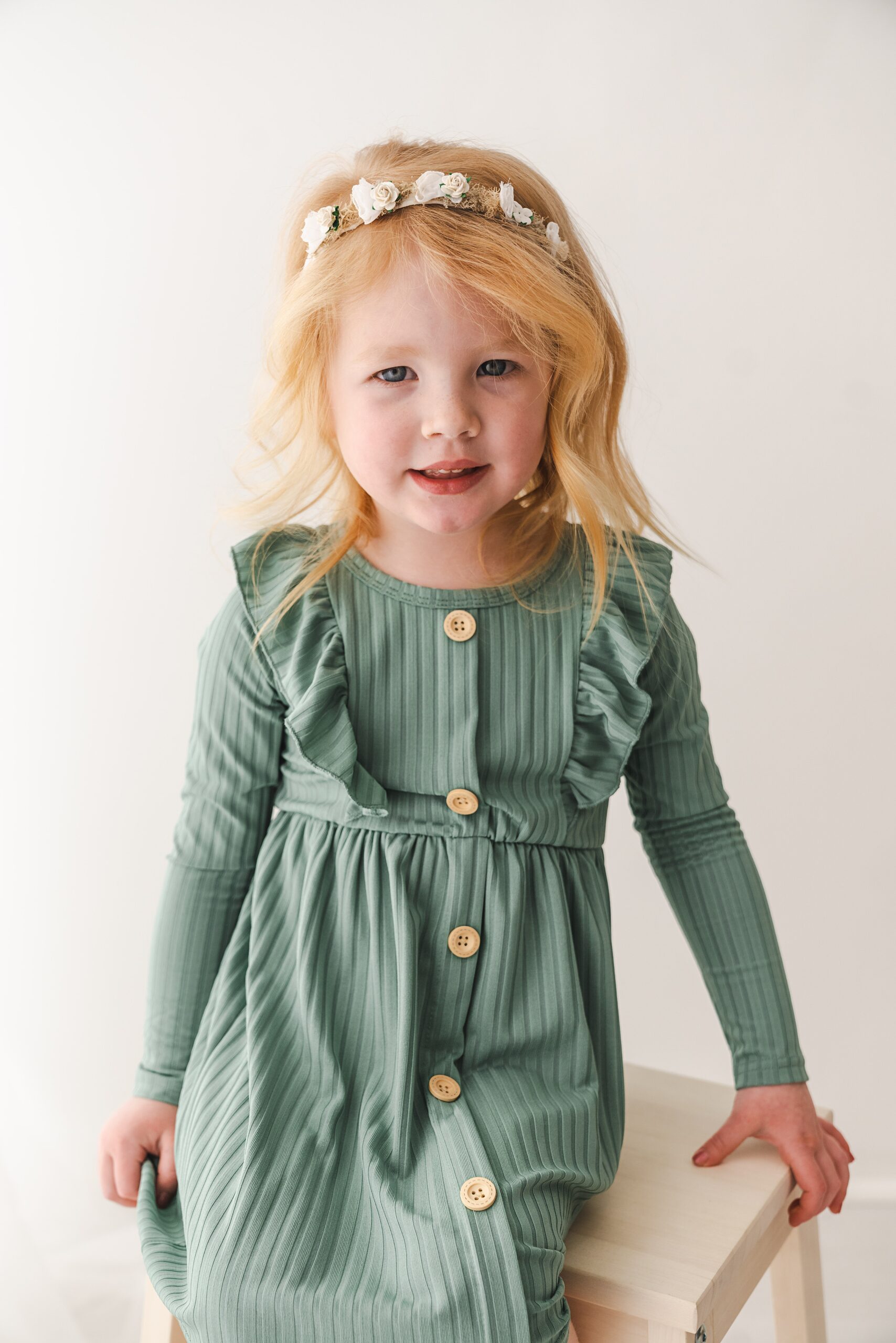 A young girl in a green dress sits on a wooden stool in a studio with a floral headband pip and pal