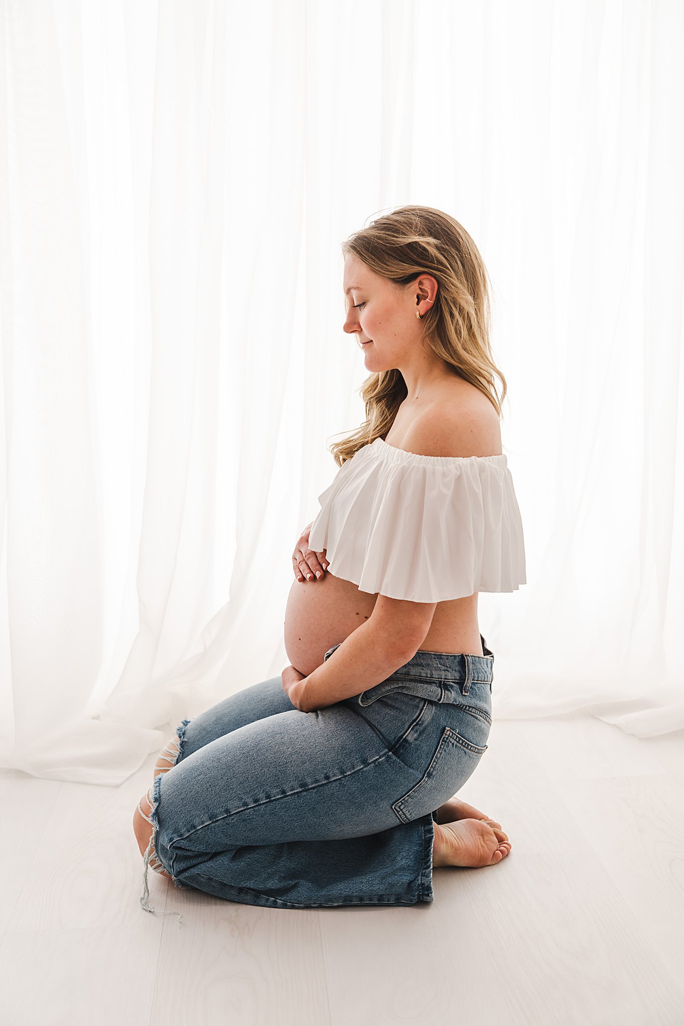 A mom to be kneels on the floor of a studio holding her exposed bump in front of a window postpartum doula minneapolis