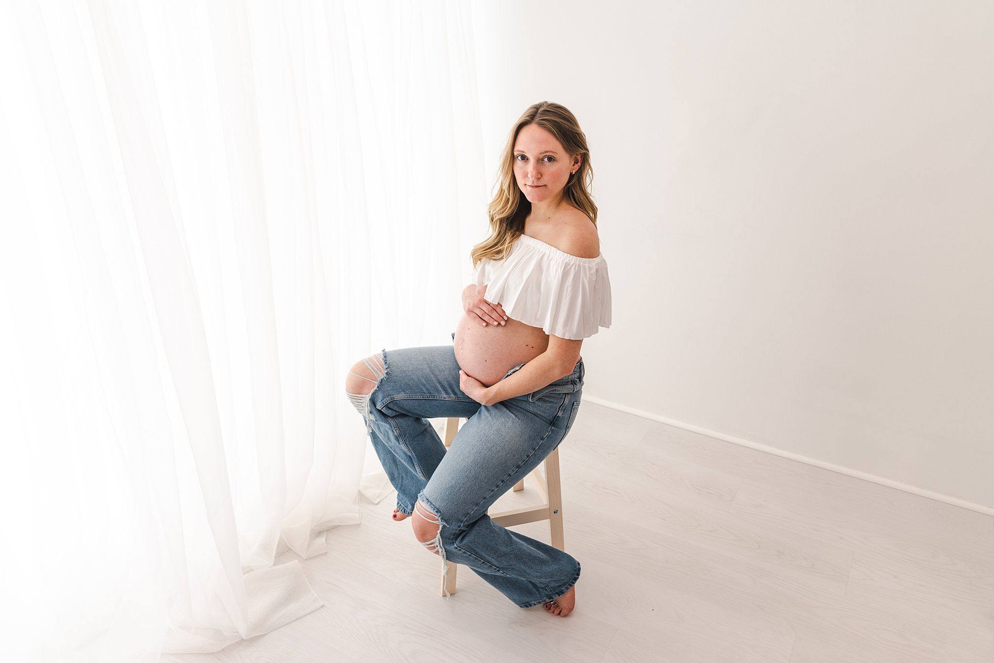 A mother to be in jeans sits on a stool in a studio holding her bump postpartum doula minneapolis