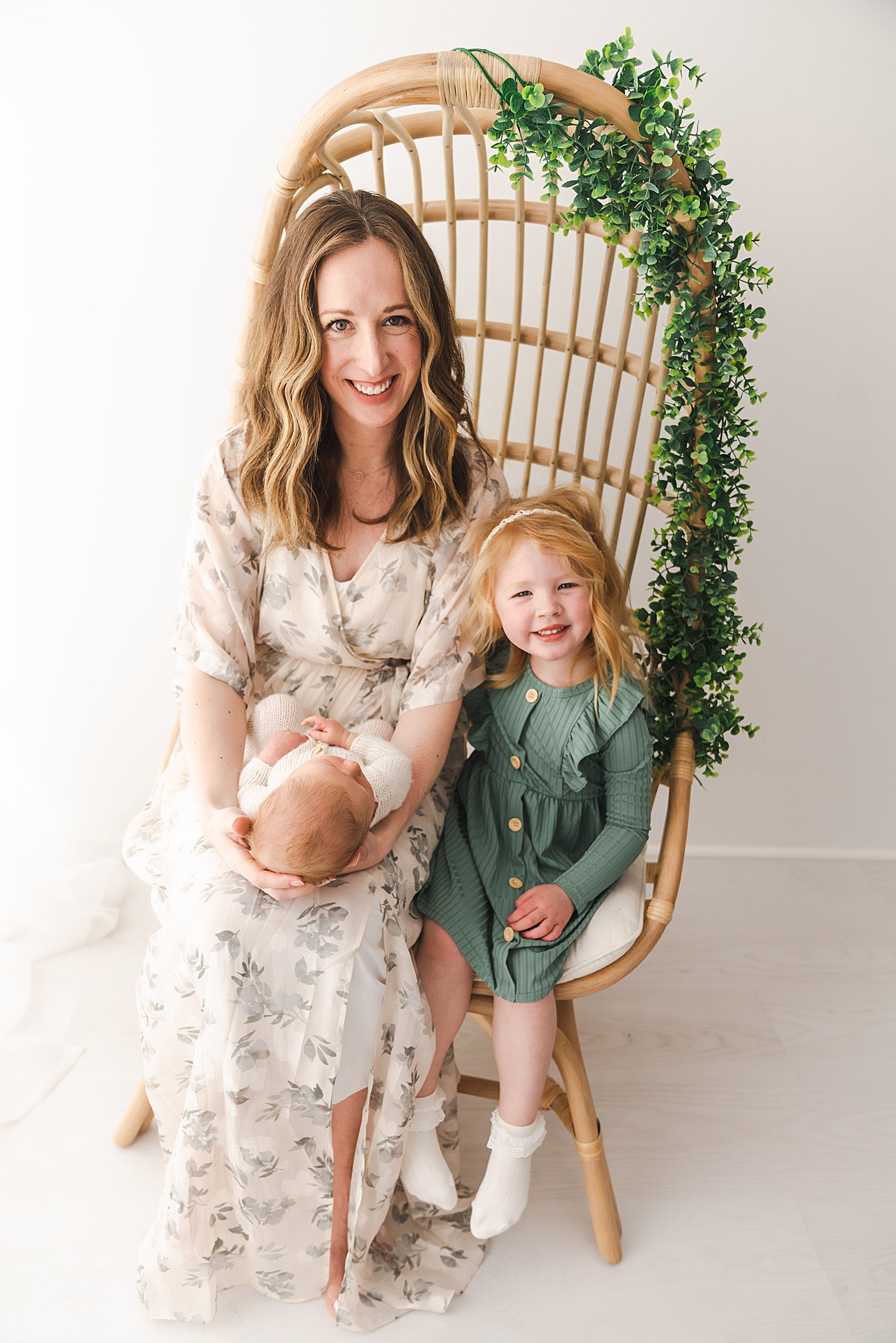 A mother and daughter sit together in a tall wicker chair in a studio while mom holds her sleeping newborn in her lap birthed mn