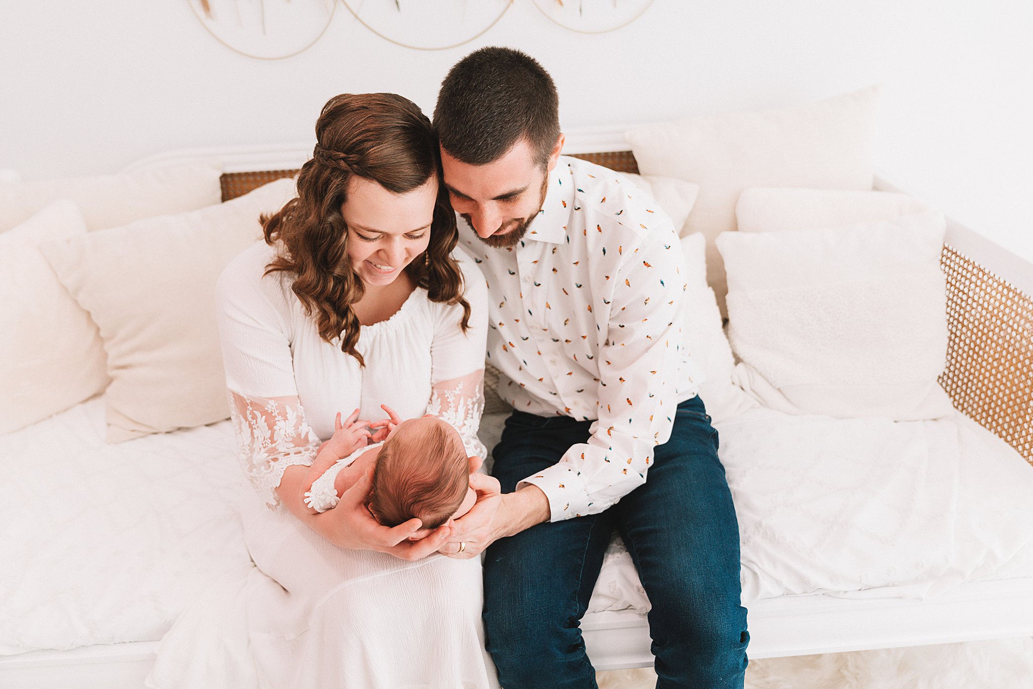 A mother and father sit on a white couch while mom looks down at her newborn baby