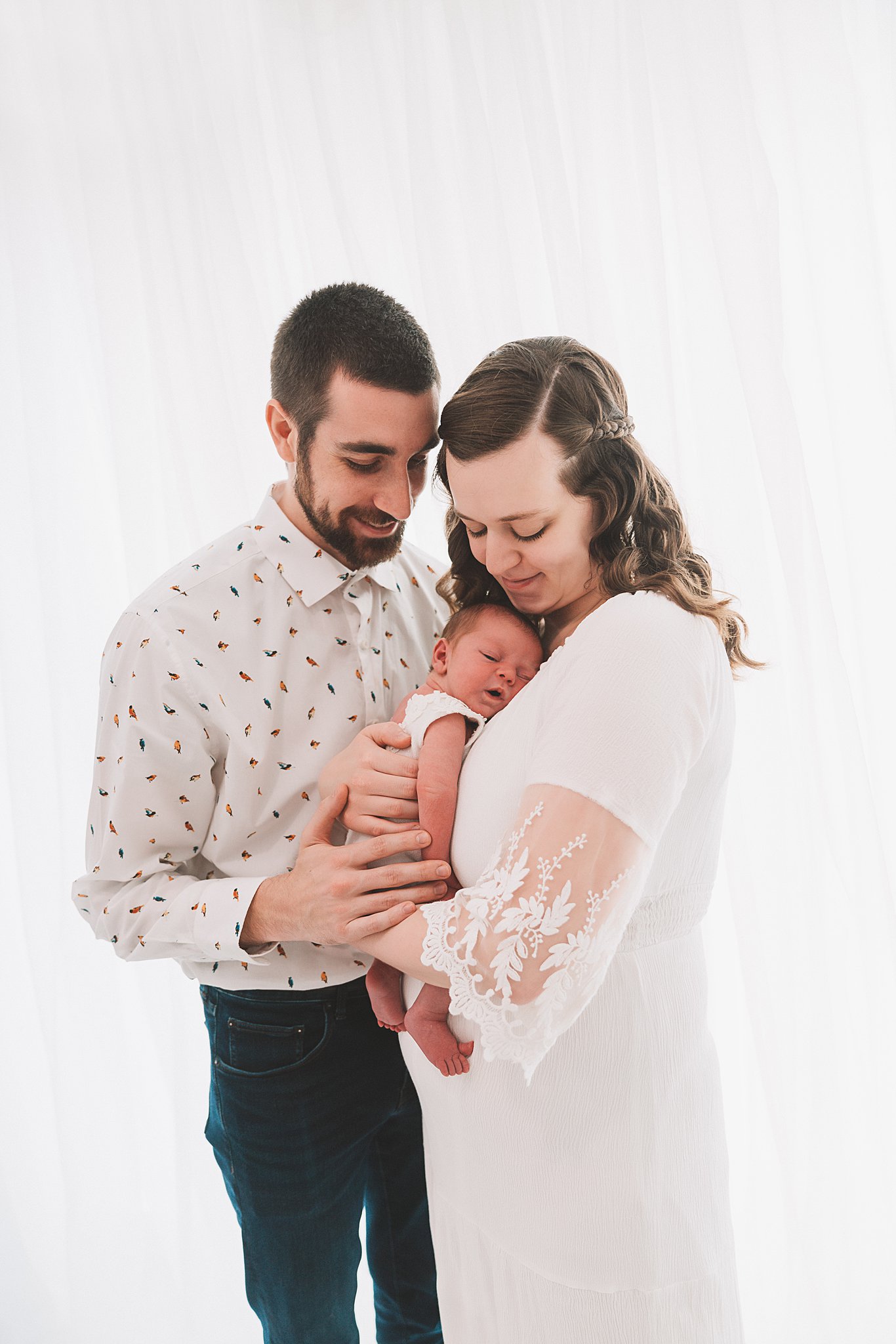 A mother cradles her sleeping baby in a white dress while dad leans in everyday miracles minneapolis