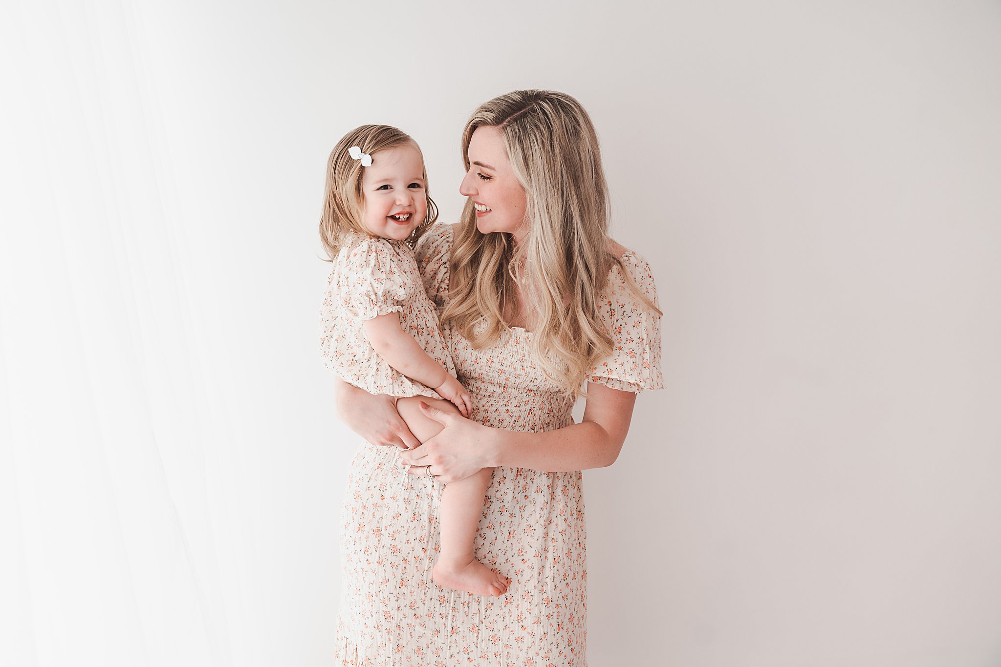 A mother in a floral pattern dress laughs and plays with her toddler daughter in a matching dress in a studio