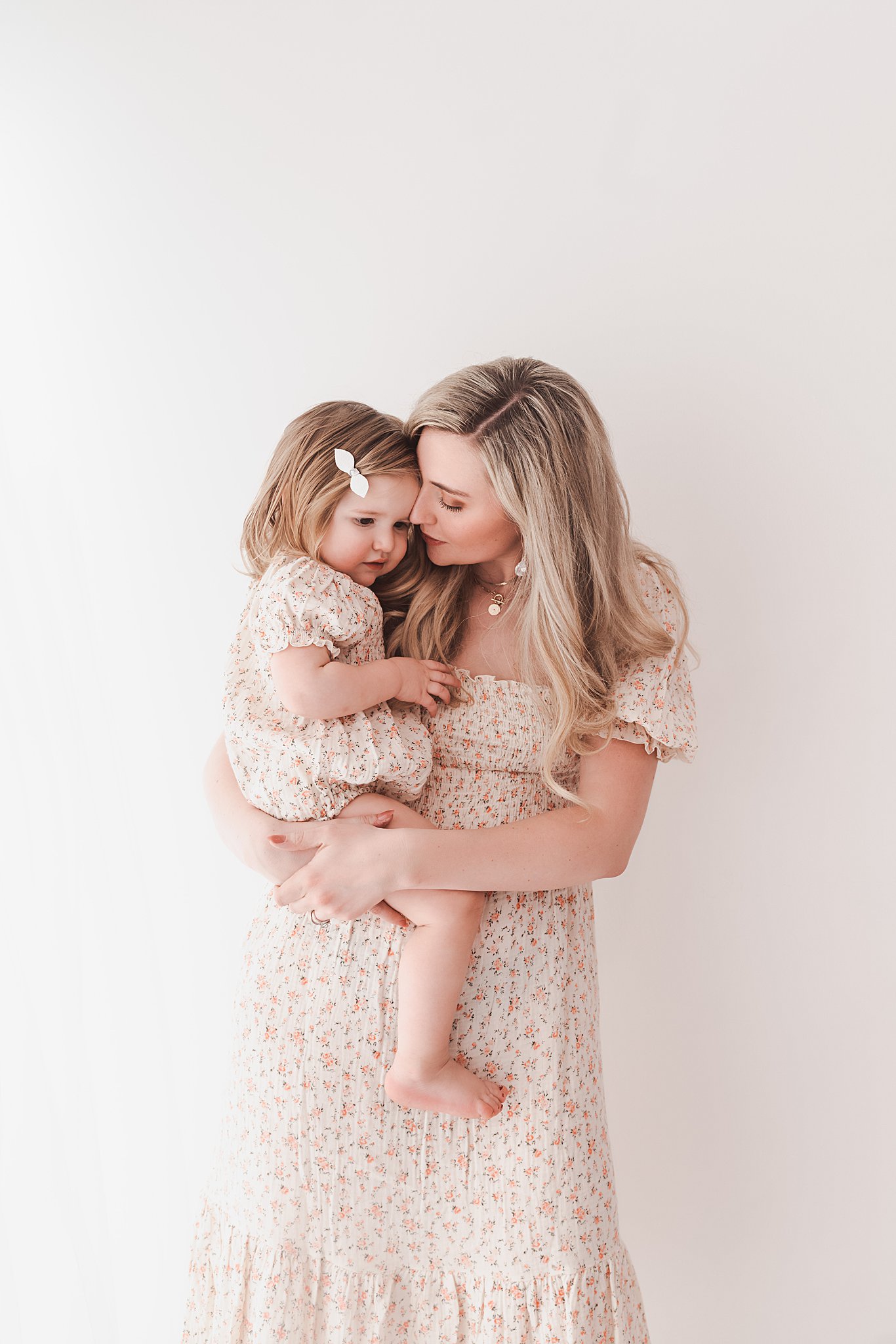 A mother in a flora pattern dress stands in a studio holding her young daughter on her him in a matching dress foss swim school minneapolis